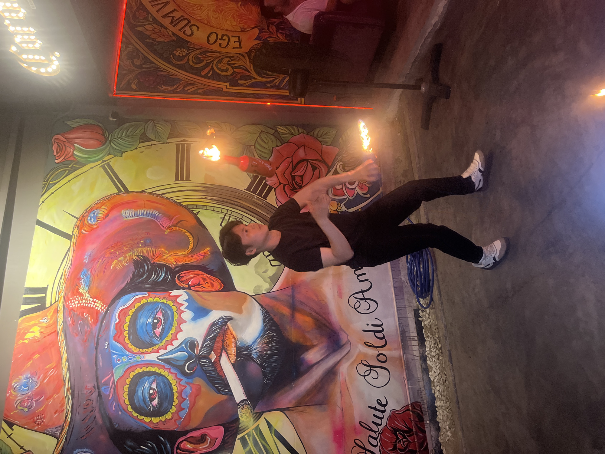 The image captures a young Asian male performing with fire in what appears to be an indoor establishment, possibly a bar or restaurant, in Đà Nẵng, Vietnam. He is dressed in a black t-shirt and black pants, wearing white and gray sneakers. The performer is actively engaged, with flames emanating from two objects he is manipulating. One flame is higher up, near his upper body, and the other is closer to the ground, near his feet.
The background is dominated by a vibrant, large-scale mural. The mural depicts a stylized portrait of a person wearing a wide-brimmed hat and elaborate face paint reminiscent of a sugar skull or Day of the Dead imagery. The mural features bold colors, including reds, blues, yellows, and purples. There are also floral elements, specifically a large red rose, and text that appears to read "Salute Soldi Am". To the right of the mural, a neon Corona sign is visible, illuminated and casting a glow. Another ornate design with the text "EGO SUM VA" is also present on the wall.
The floor is concrete and appears somewhat worn. Scattered small white pebbles are visible along the base of the wall where the mural is. A coiled blue hose lies on the ground. In the background, out of focus, other patrons can be seen sitting, with one individual wearing a white shirt. The overall lighting suggests an evening setting, with the artificial lights and the performer's fire creating a dynamic and somewhat dramatic atmosphere. The scene suggests a live performance event taking place at the venue.