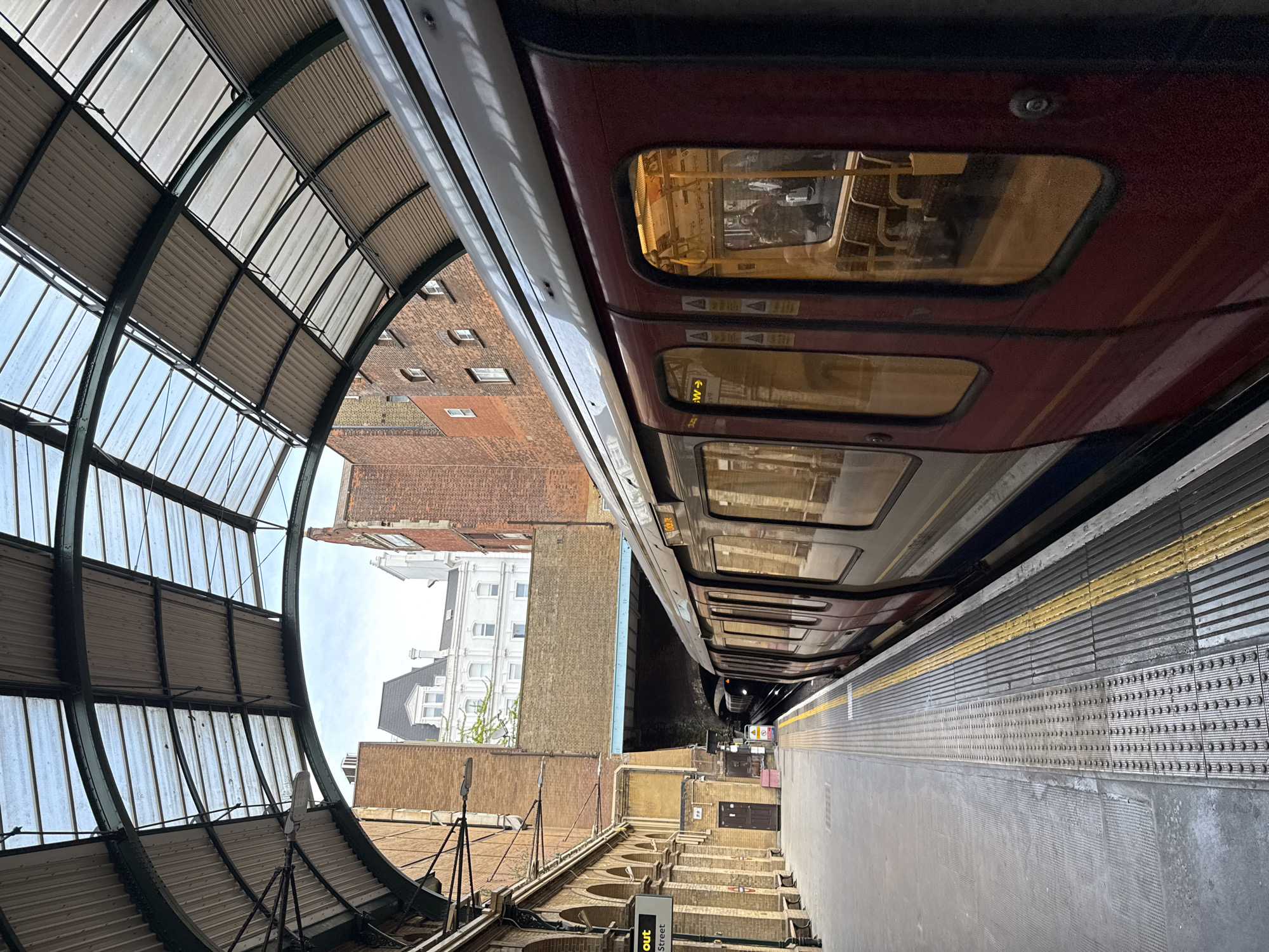The image shows a London Underground train at a station platform. The train is maroon and grey, with lit windows showing the interior. The station platform is concrete with yellow safety markings. Above the platform is a curved glass and metal canopy. Beyond the canopy are brick buildings. The scene is likely in the city of London, United Kingdom.