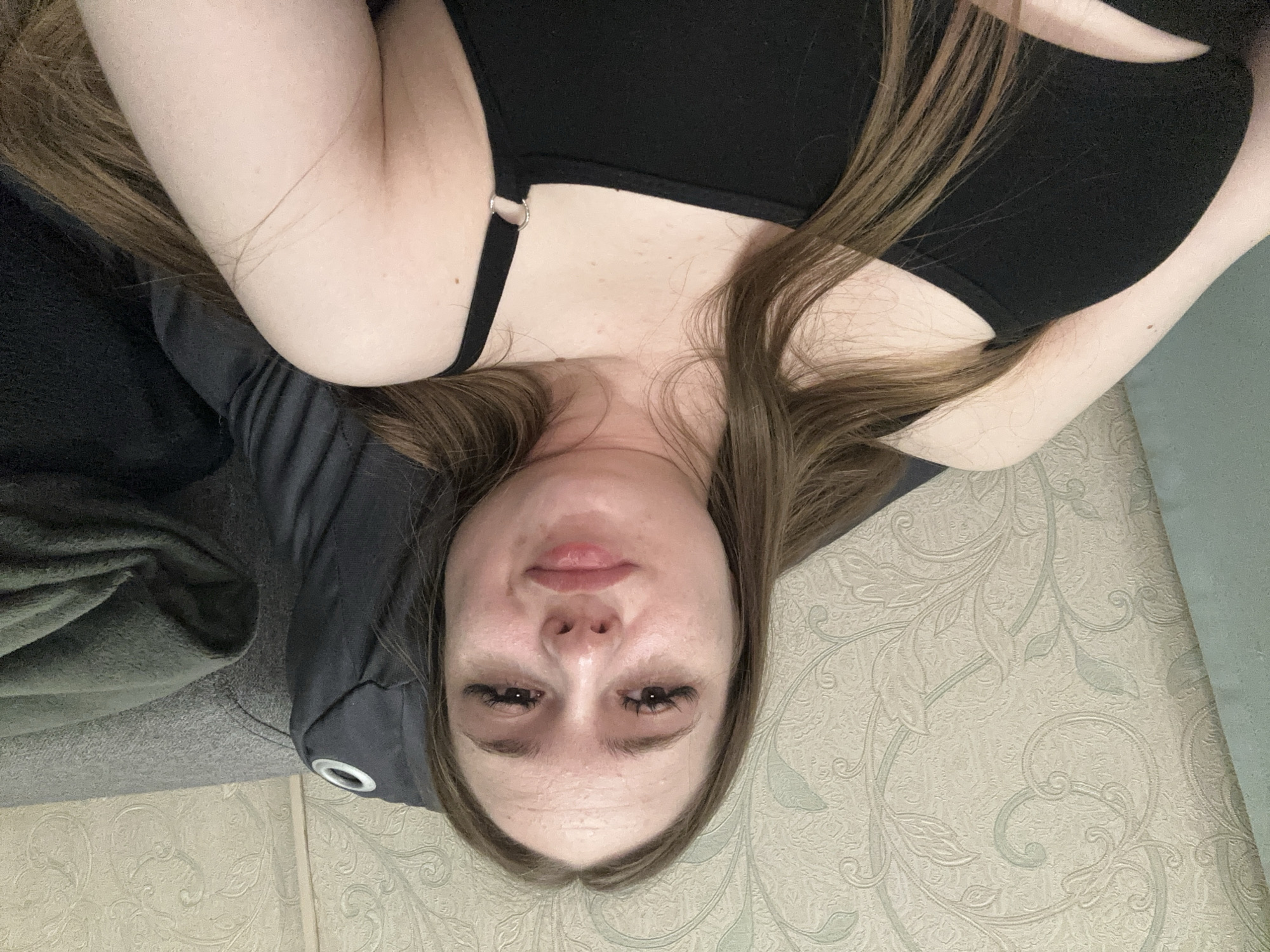 A close-up, high-angle selfie captures a young Caucasian woman from the chest up, lying down and looking directly at the camera. She has long, light brown hair that is partially covering her shoulders and chest. Her skin is pale with some freckles visible on her chest and neck. She is wearing a black tank top with a thin strap and a silver ring detail. Her makeup includes dark, dramatic false eyelashes and a light pink lipstick. The background consists of textured wallpaper with a light floral pattern in neutral tones, and a grey sofa. A dark green blanket is folded on the sofa to the right of the woman. The lighting is soft and diffuse, creating subtle shadows.