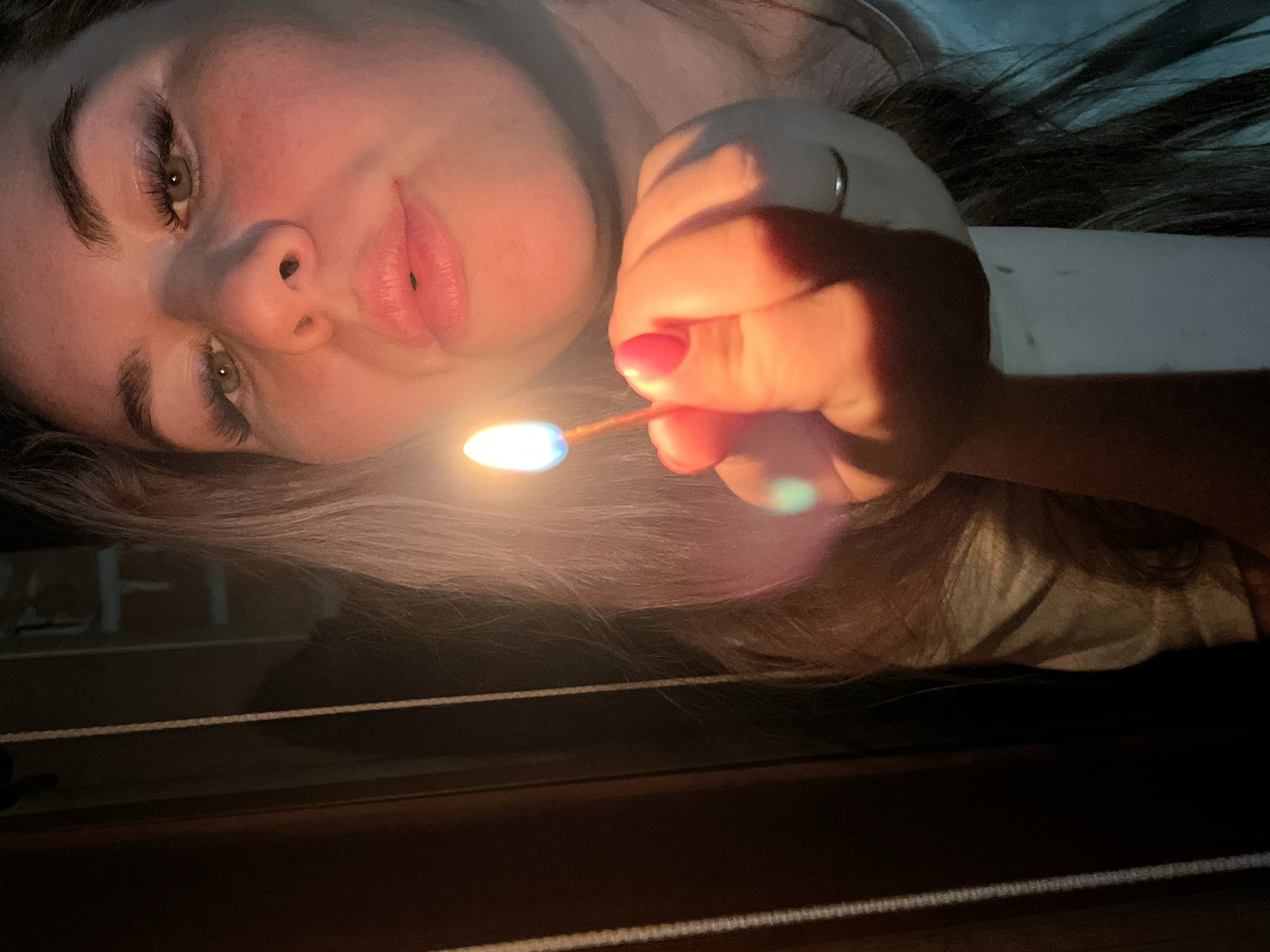 A woman with fair skin, dark eyebrows, and green eyes lies on her side, holding a lit match. Her nails are painted red. She is likely indoors; part of a table is visible in the foreground. The city of Chaikovsky, Russia, cannot be determined from the provided image.