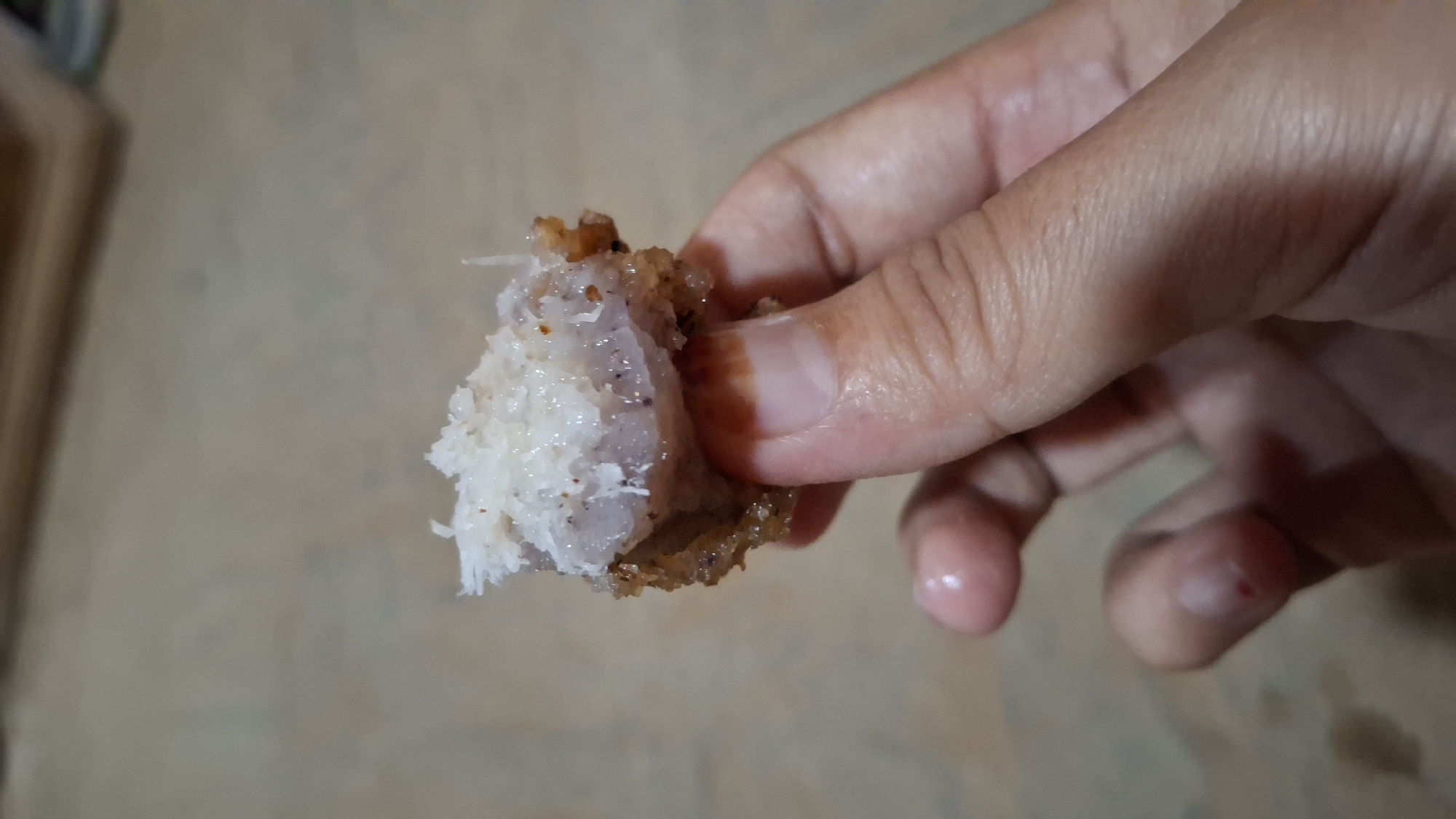 A close-up shot shows a person's hand holding a piece of fried food that has been coated in shredded coconut. The food item is golden brown and appears to be slightly crispy on the outside. The inside is lighter in color and has a moist, possibly starchy texture. The shredded coconut is white and finely grated, clinging to the surface of the food. The background is blurred and appears to be a neutral-colored surface, possibly a table or floor. The lighting is soft, highlighting the details of the food and the hand.