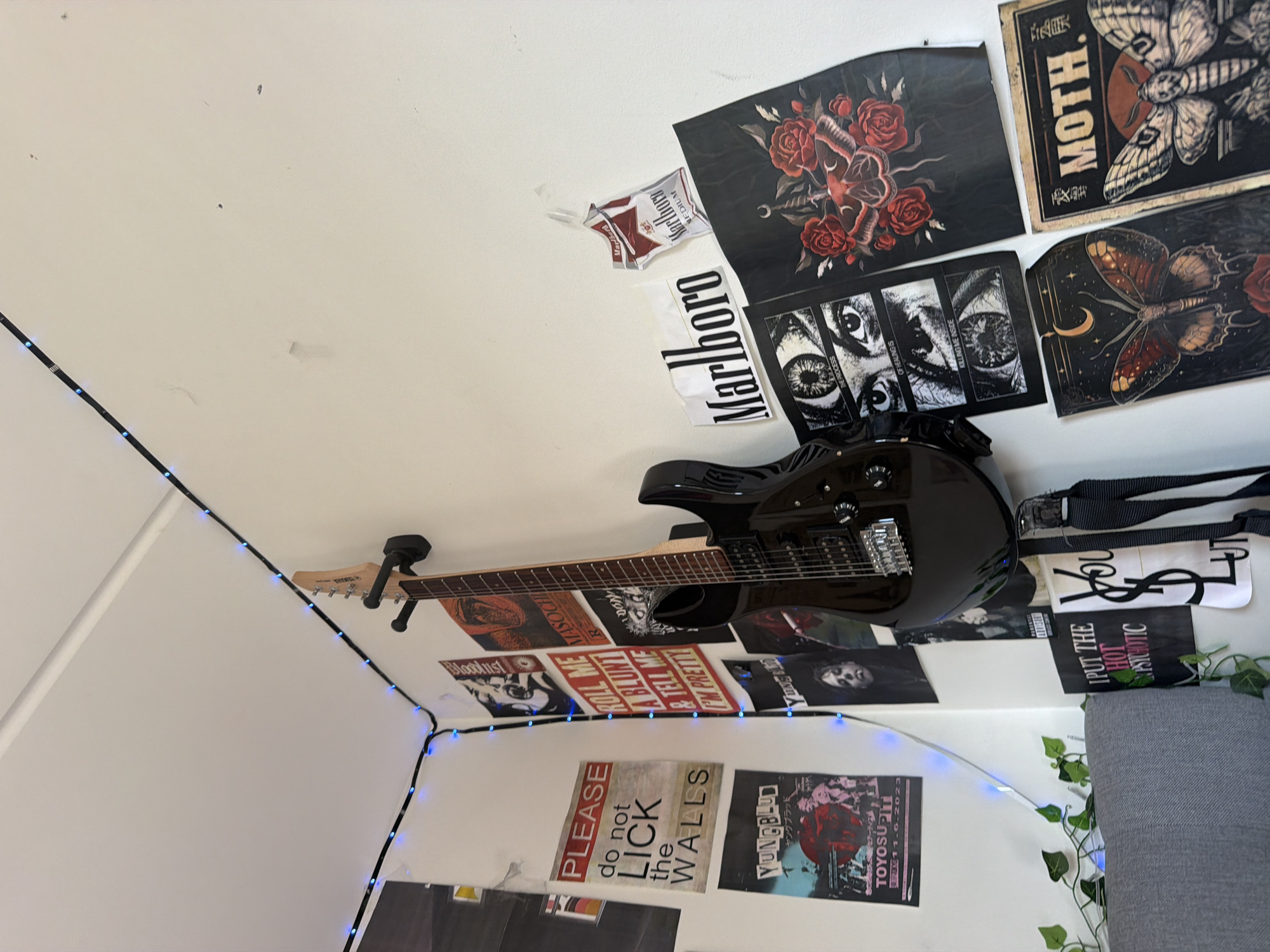 The image shows a corner of a room decorated with band posters and a black electric guitar mounted on the wall. Blue string lights are draped along the top corner of the walls. Posters visible include "PLEASE do not LICK THE WALLS," "Yungblud," "ROLL ME A BLUNT & TELL ME I'M PRETTY," and posters with images of eyes, moths, and roses. There is also a Marlboro poster and what appears to be a cigarette pack. A portion of a grey sofa is visible in the lower left corner, with some artificial greenery draped over it. The wall appears to be painted white and has some scuff marks. The scene is indoors and does not provide any information that would place it in Oslo, Norway.