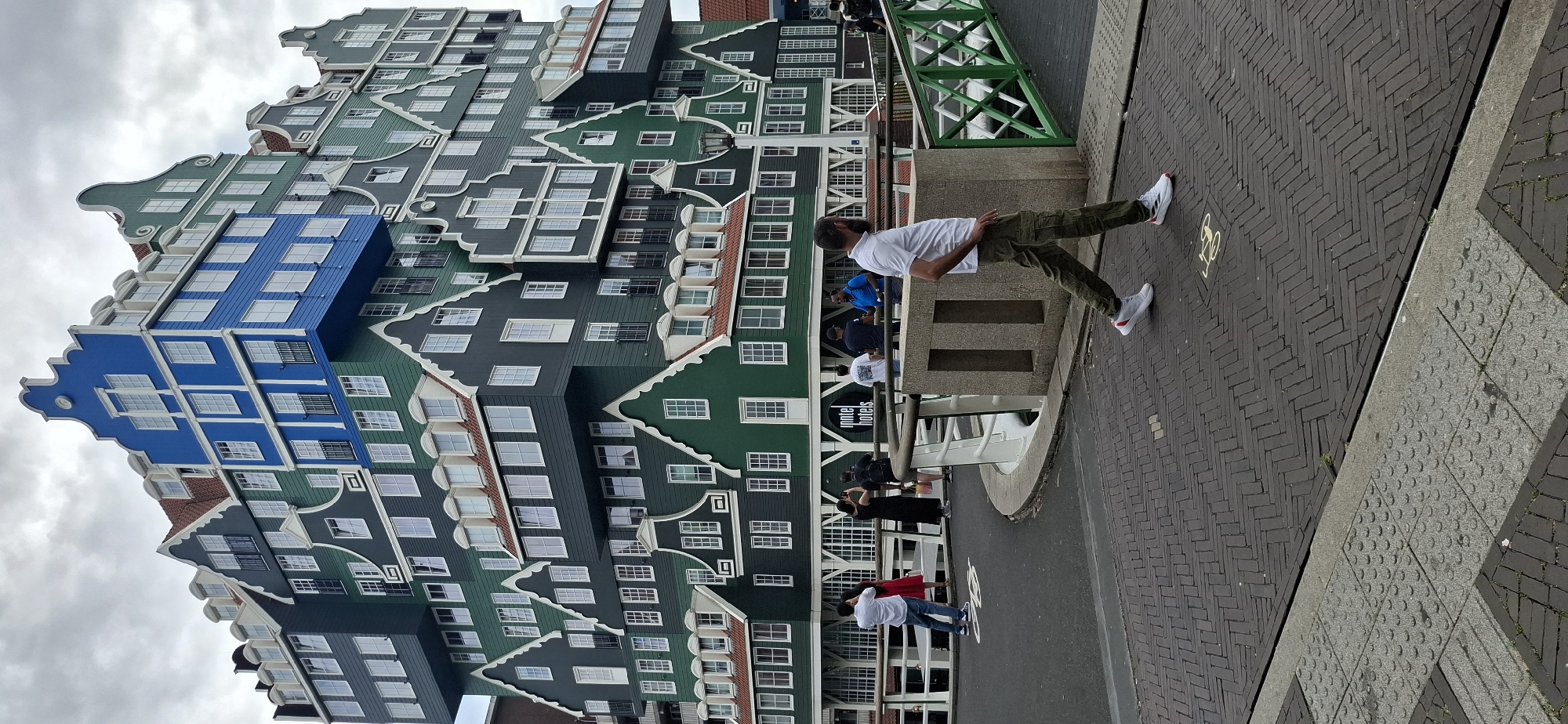 The image shows a man walking across a bridge in front of the Inntel Hotels building in Zaandam, Netherlands. The building is a distinctive postmodern structure designed to resemble a stack of traditional Dutch houses. The man is wearing a white polo shirt, olive green cargo pants, and white sneakers. He is walking from right to left in the frame.
In the background, several people are standing on the bridge, some of whom are taking photos of the hotel. The bridge has white railings and green decorative elements. The foreground of the image is a brick-paved walkway, with a section of tactile paving for visually impaired pedestrians. The sky is overcast.