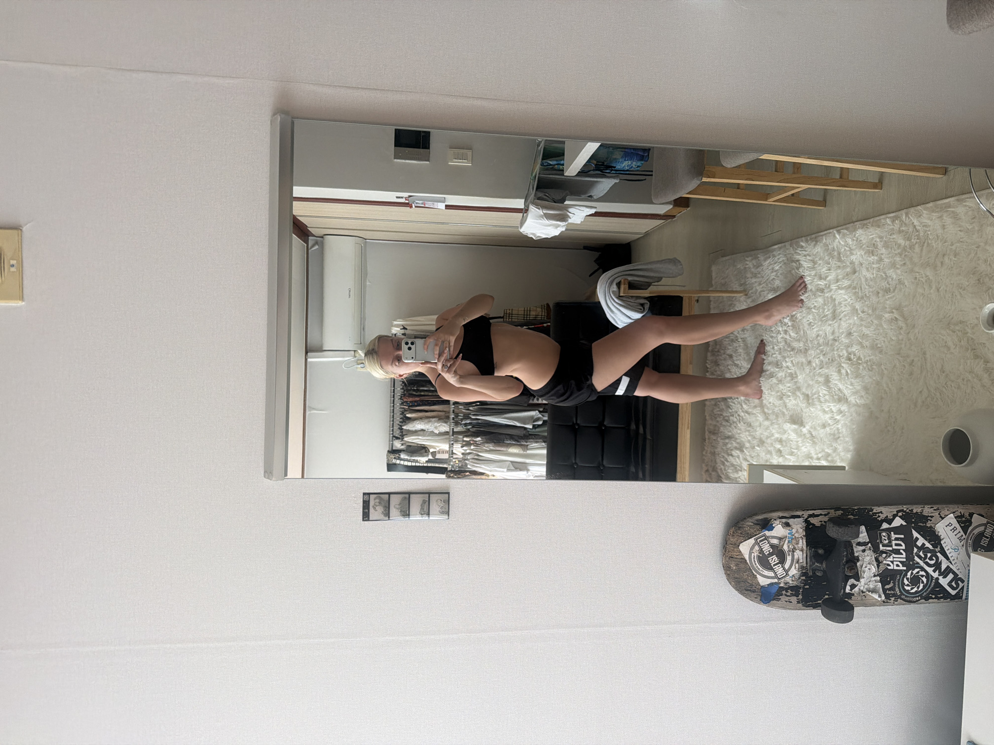 The image depicts a woman in athletic attire taking a mirror selfie inside an apartment in Seoul, South Korea. The apartment features white walls, a wardrobe with hanging clothes, a white shag rug, and a skateboard leaning against the wall. The woman is wearing a black sports bra and black shorts, and she is holding a smartphone.