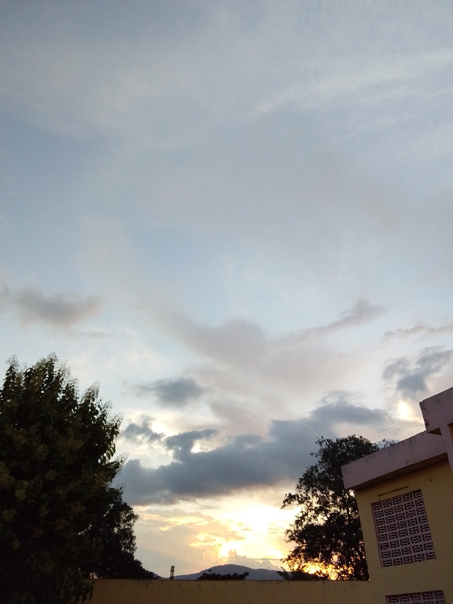The image depicts a sunset scene in Serti, Nigeria, with a partly cloudy sky casting a soft glow over the landscape. In the foreground, a building with a yellow facade and a decorative concrete block window is visible on the right. A dark, leafy tree stands to the left of the building, partially obscuring the sky. The horizon is lined with rolling hills. The overall impression is one of a peaceful evening in a semi-urban or rural setting.