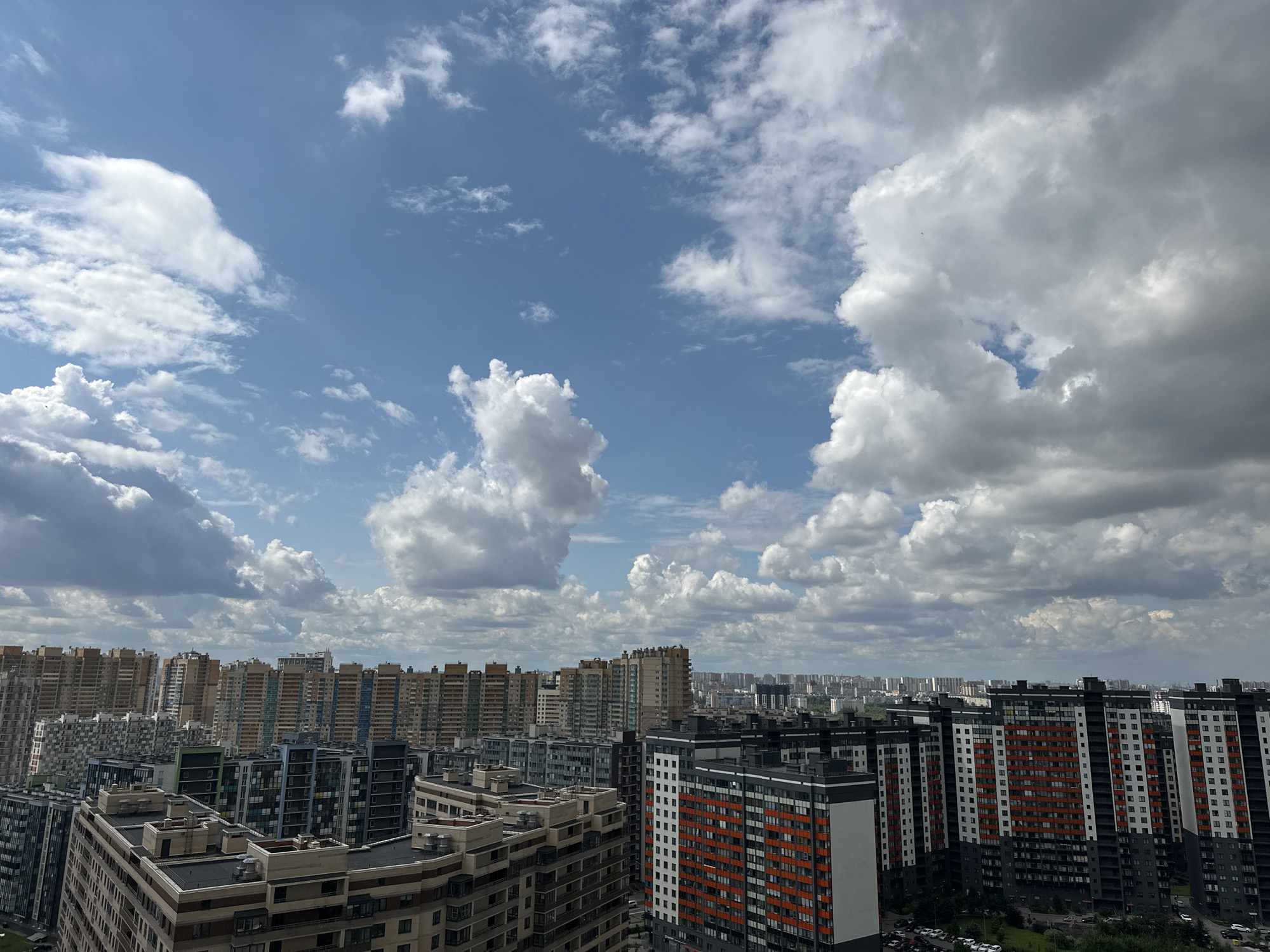 This is a high-angle, eye-level, daytime, medium shot of a cityscape in Dnp Ryzhiki, Russia. The sky is a vibrant blue, dotted with fluffy white clouds, some of which appear darker and heavier, suggesting impending rain. In the foreground, a cluster of modern, multi-story apartment buildings dominates the scene. These buildings feature a mix of architectural styles and colors, with prominent use of white, gray, and orange facades. Many of the buildings have numerous windows and balconies, indicating residential use. The buildings are densely packed, creating a sprawling urban landscape. In the mid-ground and background, more apartment buildings and structures extend into the distance, forming a vast, urban horizon. Some of these distant buildings are lighter in color, appearing as a haze against the sky. The scene is devoid of people, cars, or any immediate signs of human activity, emphasizing the vastness of the urban development and the immensity of the sky above.