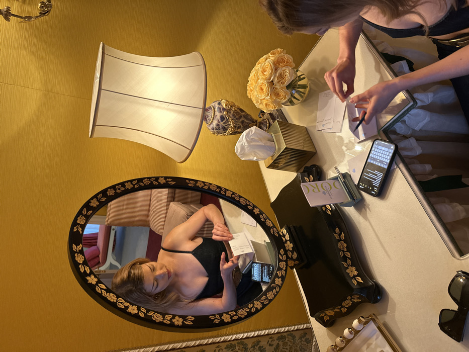 A top-down view captures a woman writing at a vanity table. A bouquet of light yellow roses sits in a clear vase. A gold tissue box, a DORO-branded notepad, and a phone displaying a keyboard lie nearby. The woman's reflection is visible in a dark, oval mirror decorated with gold flowers. A large lampshade stands beside the mirror on a yellow wall. Sunglasses rest on the vanity's white surface.