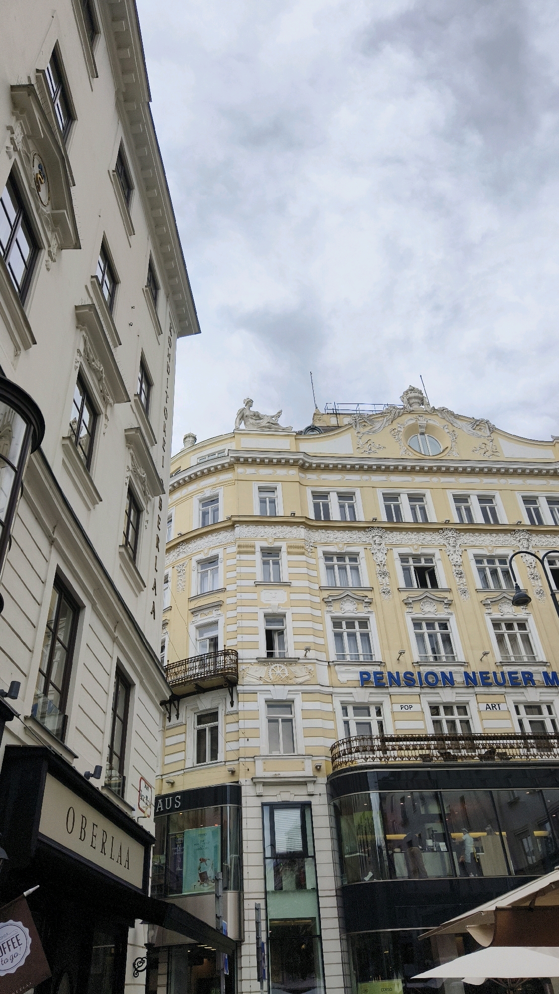 The image depicts a street view in Vienna, Austria, focusing on the architecture of buildings. In the foreground, a white building with ornate detailing and dark-framed windows is on the left. To its right, a light yellow building with a similar architectural style, featuring multiple windows and decorative elements, is prominent. A sign on the yellow building reads "PENSION NEUER M". The sky above is overcast with clouds. The street level appears to have storefronts, including one labeled "OBERLAA" on the white building.