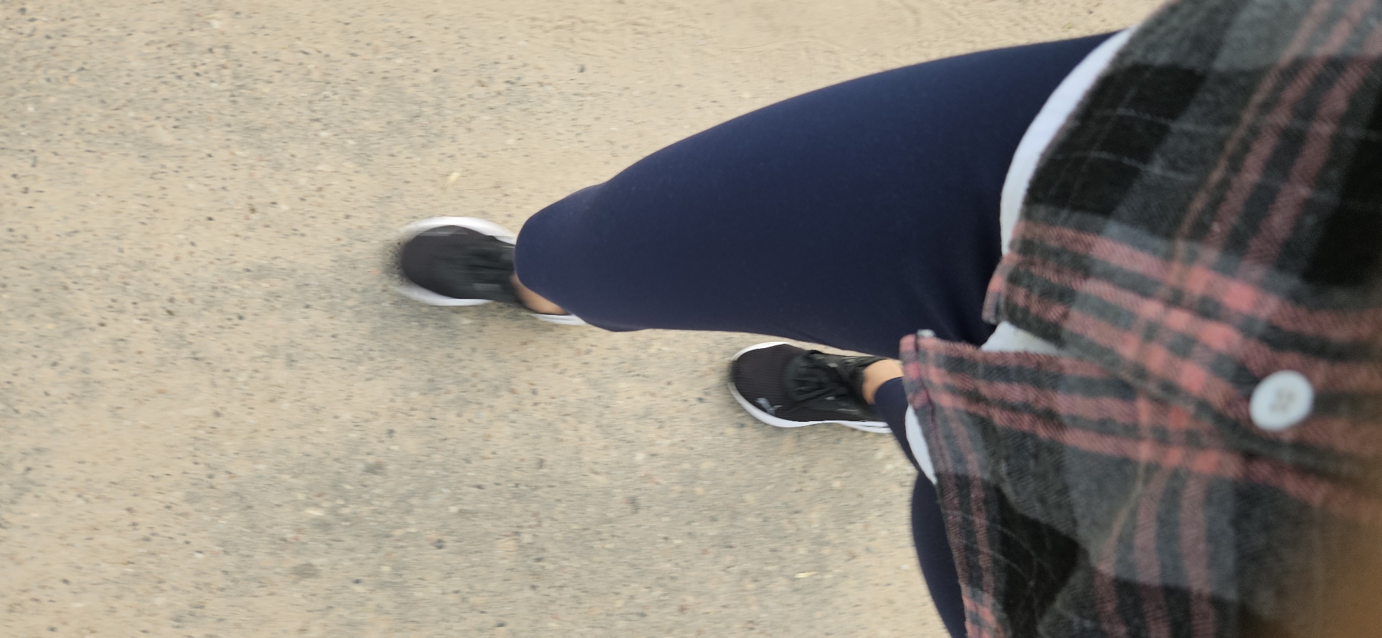 A first-person perspective shot shows a person walking on a light-colored, gritty surface, likely a sidewalk or path. The individual is wearing dark blue leggings, black and white athletic shoes, and a plaid shirt with pink and gray stripes. The lower part of the legs and feet are in focus, while the upper body and surrounding environment are blurred due to motion. The camera angle is looking down at the person's legs as they walk. There is no discernible location within the city of Vitsebsk, Belarus, as the background is generic.