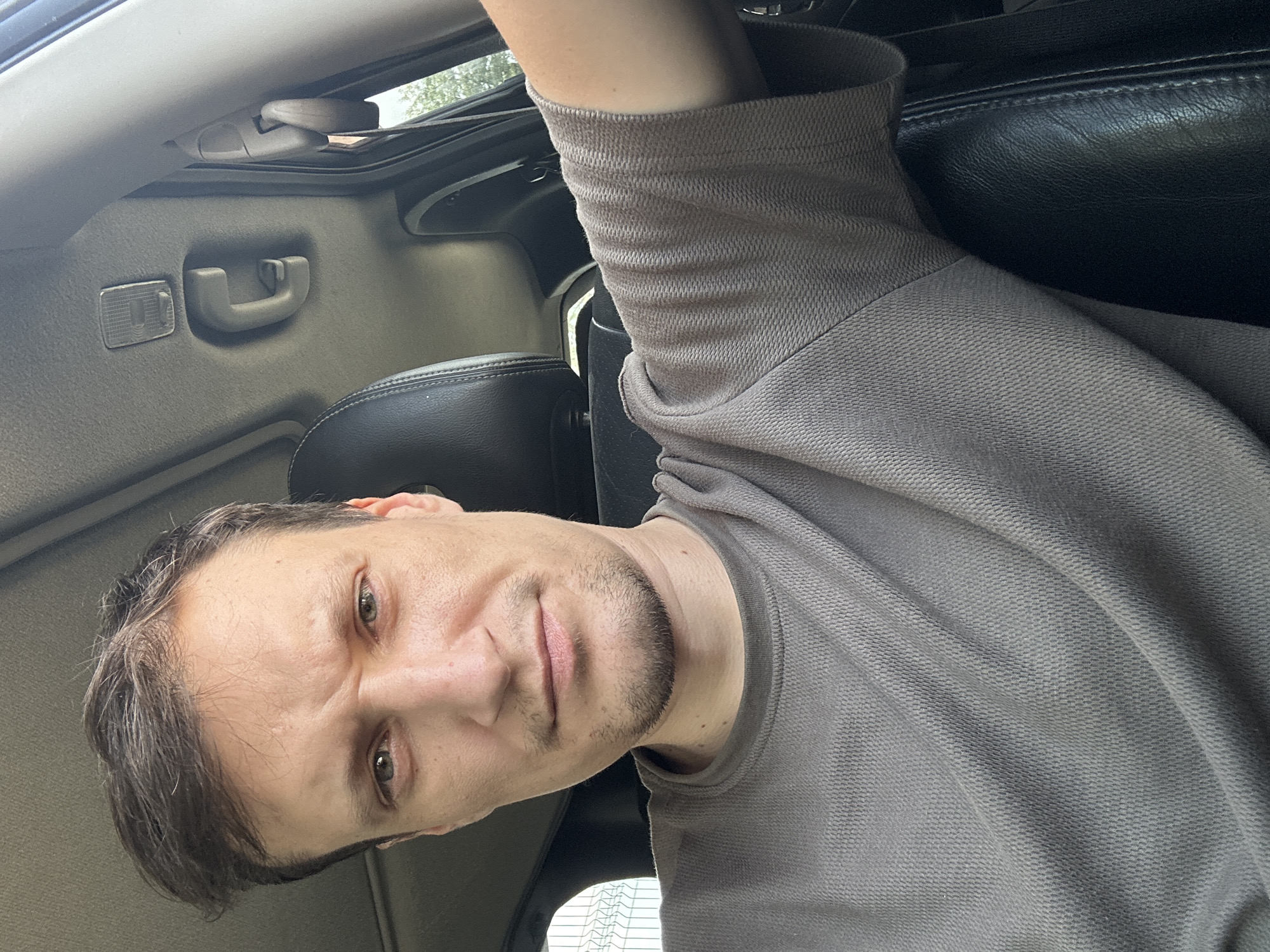 A man is captured in a selfie from the chest up, seated inside a vehicle. The man has short, dark brown hair, light green eyes, and a light beard. He is wearing a grey, short-sleeved t-shirt. The interior of the car is visible, including the headrest of a black leather seat, the car's ceiling with a grab handle and a light fixture, and the window. The man is looking directly at the camera with a neutral expression. The image was taken in Shatava, Ukraine.