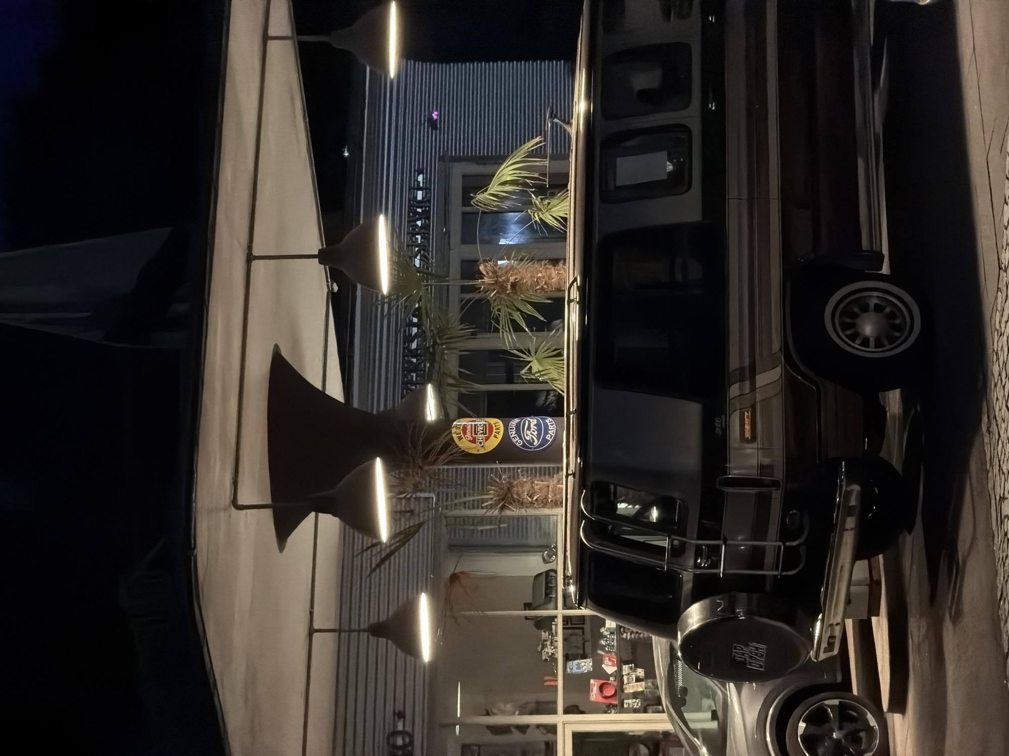 The image shows an outdoor scene at night, likely a service station or auto repair shop, illuminated by hanging lamps. In the foreground, a dark-colored van with a spare tire mounted on the rear is parked. To the left of the van, a gray car is partially visible. The background features a building with large windows, displaying shelves with various items. Behind the building, there are palm trees and signage, including a round sign with "CHEVROLET" and "Ford PARTS" logos. The overall impression is of a retro or classic automotive-themed establishment. The location is identified as Garmisch-Partenkirchen, Germany, suggesting a specific context for this scene, possibly a unique business or attraction within the town. There are no people visible in the image.