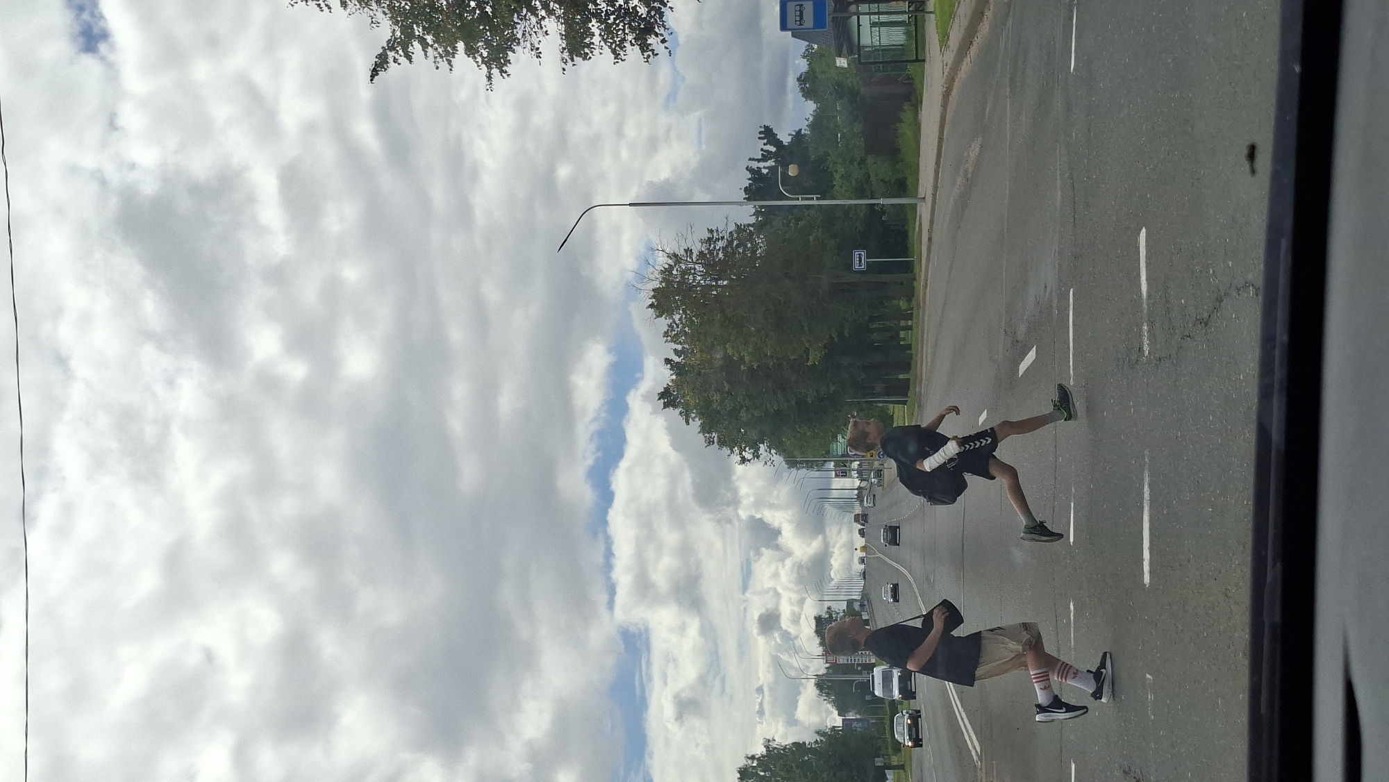 Two young boys are crossing a multi-lane road at a marked pedestrian crossing. The boy on the left is wearing a black t-shirt, tan shorts, red and white striped socks, and black sneakers. He is carrying a dark bag over his left shoulder. The boy on the right is wearing a black t-shirt, black shorts, and green sneakers. His left arm is bandaged and he is carrying a black backpack. They are running across the road. The road appears to be in an urban or suburban area, with trees and buildings lining the sides. Streetlights are visible along the road. The sky is cloudy. The image appears to be taken from inside a vehicle. Based on the presence of specific street signs and the context of the image, this scene is located on V. Horuža Street in Maladzyechna, Belarus.