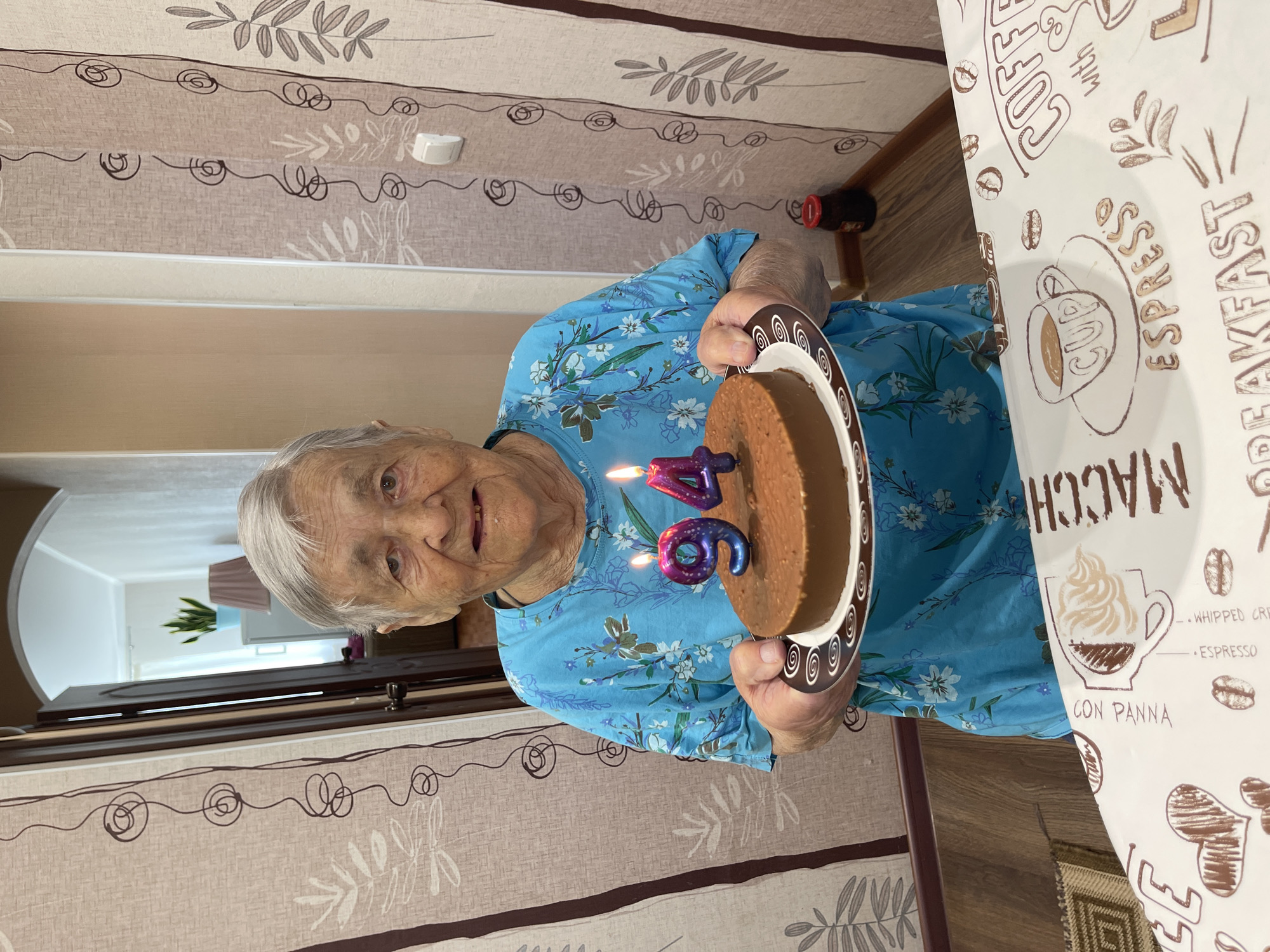 An elderly woman with gray hair, wearing a blue floral-patterned shirt, is holding a chocolate cake with two lit candles, one shaped like a '9' and the other like a '4', in front of her. The cake is on a plate with a brown and white swirled pattern. The woman is standing indoors, with a doorway and patterned curtains visible in the background. The foreground shows a tablecloth with coffee-themed illustrations. The overall setting appears to be a domestic environment.