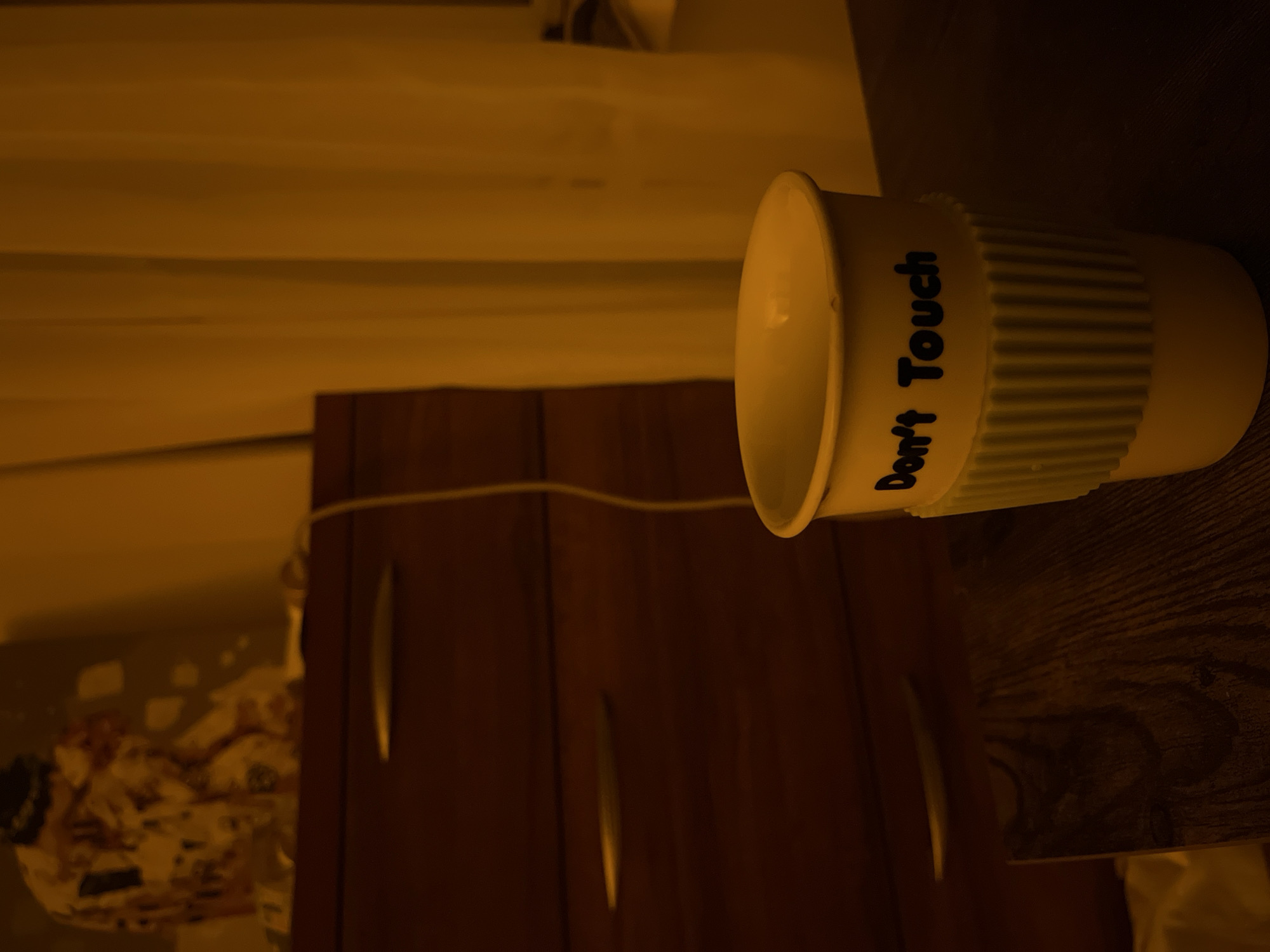 A close-up, low-angle shot captures a white paper cup with a blue ribbed sleeve and black text that reads "Don't Touch" in the foreground. The cup sits on a dark wooden surface, possibly a table, with a blurred background. To the left, a dark brown dresser with three drawers, each featuring a silver handle, is visible. Above the dresser, a blurry object with a patterned fabric, possibly clothing or a stuffed animal, is placed. To the right of the cup, a sheer, yellow curtain hangs, obscuring the view beyond. The lighting in the scene is warm and casts long shadows, suggesting an indoor setting possibly at night or in a dimly lit room. The overall ambiance is one of a personal space, with the "Don't Touch" message on the cup adding a potentially playful or cautionary element.