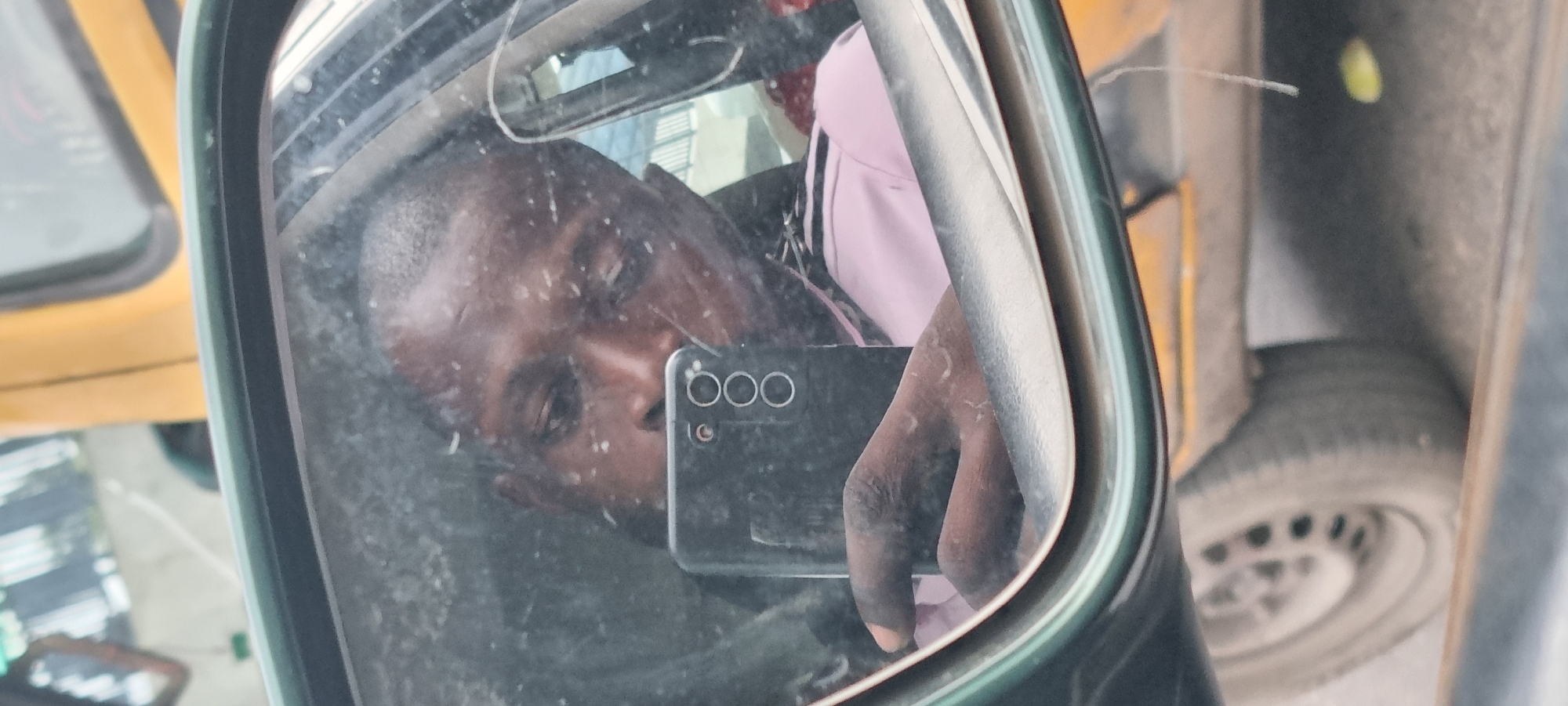 The image displays a close-up view of a person's reflection in a vehicle's side mirror. The person, who has dark skin and short, dark hair, is holding a black smartphone with a triple camera array visible on its back. The reflection captures the person's face in profile, looking slightly to the left, seemingly at the phone's camera to take a picture or video. The hand holding the phone is visible towards the bottom right of the reflection.
The surface of the mirror appears somewhat dusty or smudged, with numerous specks and streaks slightly obscuring the clarity of the reflection. Beyond the mirror, to the left, parts of a yellow vehicle or structure are visible, including a window frame. To the right, a vehicle's tire and wheel rim are discernible. A hint of a pink garment or accessory is also reflected behind the person's head. The overall lighting suggests it is daytime. The setting appears to be an outdoor, possibly urban environment, consistent with the streets of Lagos, Nigeria. There is no visible text.