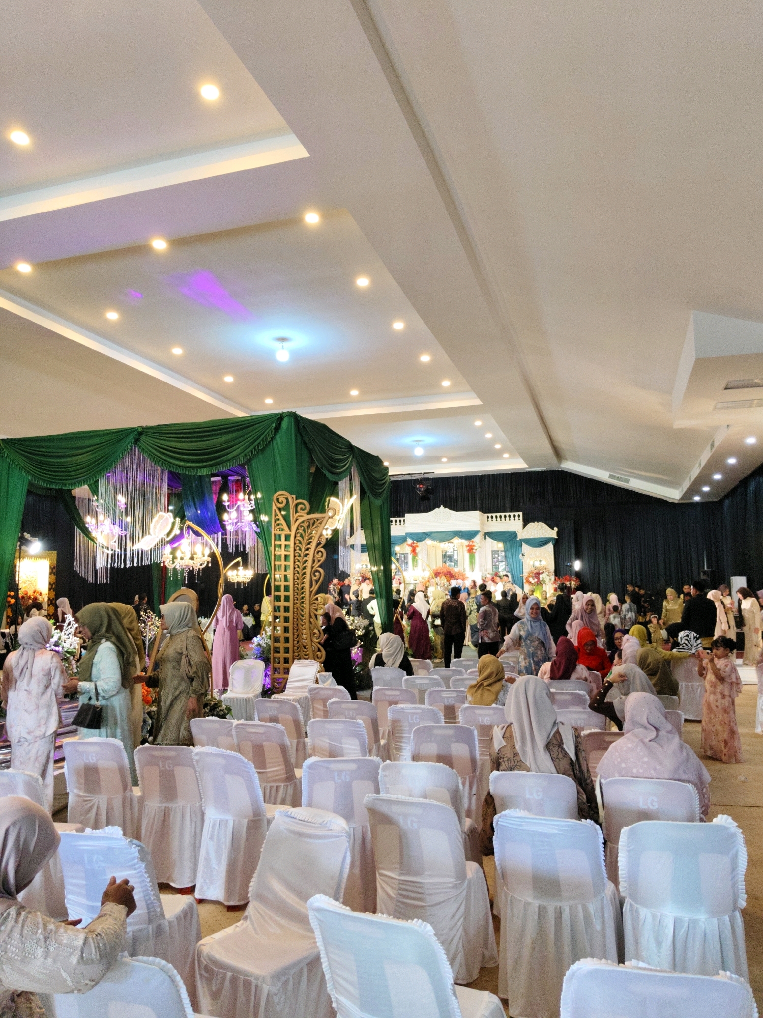 A wedding reception is taking place in a large indoor venue. The room is decorated with green drapes and ornate gold structures. White chairs are set up in rows facing a decorated stage area. Numerous people, mostly women wearing headscarves, are mingling and seated. The lighting appears to be a mix of warm and cool artificial lights, with some purple uplighting visible on the ceiling. Based on typical wedding venue decorations and the attire of the attendees, this scene is likely from a wedding ceremony or reception in Lhokseumawe, Indonesia.