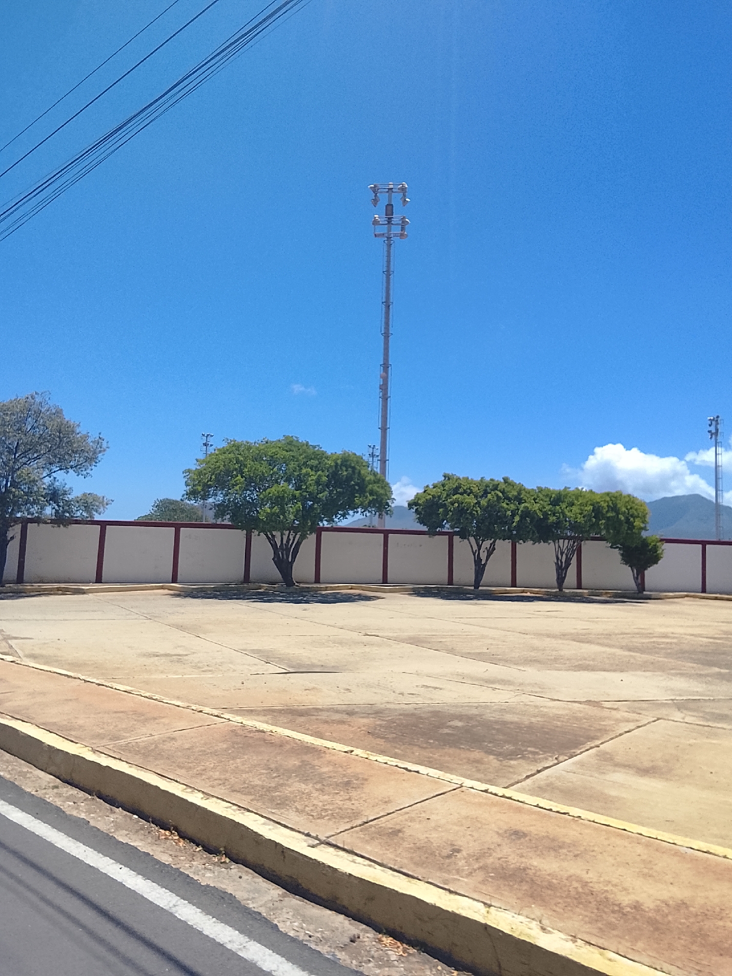 This image presents an outdoor scene under a bright, clear blue sky on a sunny day. In the foreground, a gray road with a white painted line transitions into a yellow-painted concrete curb, which borders a wide, stained concrete paved area.
Stretching across the midground is a low, white wall punctuated by several reddish-brown vertical supports. Behind this wall, a line of trees is visible. The trees on the right have dense green foliage, while the one on the far left appears to have lighter, possibly drier, leaves.
In the background, a tall, slender metal communication tower stands prominently to the right of center. Another similar tower is partially visible further to the right. Distant mountains rise on the horizon on the right side, partially shrouded by white clouds. Multiple dark power lines cut diagonally across the upper left portion of the bright sky.
There are no people or discernible activities taking place. The scene appears still and functional. The strong sunlight indicates it is daytime, likely morning or early afternoon. No text is visible anywhere in the image. The setting, with its specific vegetation, infrastructure, and bright conditions, aligns with a location such as Juan Griego, Venezuela.