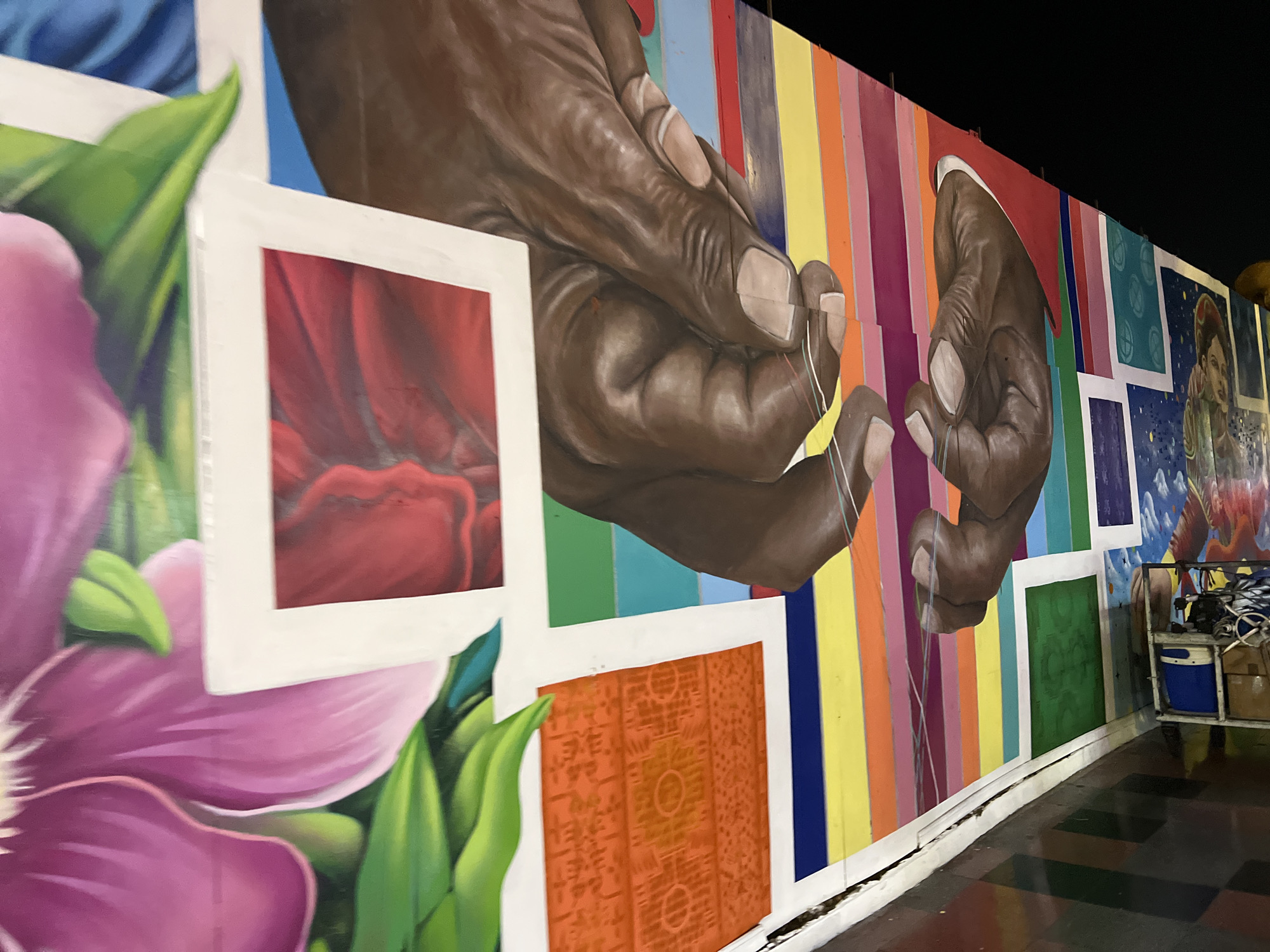 The image shows a section of a vibrant mural on a wall in Arica, Chile. The mural features large, detailed paintings of hands with dark skin, appearing to be engaged in some activity, possibly playing a stringed instrument, as thin colorful threads are visible between the fingers. These hands are positioned against a backdrop of vertical, brightly colored stripes in shades of pink, yellow, orange, green, blue, and purple. To the left of the hands, there are painted representations of large pink and red flowers with green leaves, framed by white squares. Further to the right, there are additional painted panels, one with a blue background and a pattern of white circles, and another with a dark sky, stars, and a depiction of a person. In the lower right corner, a metal cart with wheels is visible, holding various items, including a blue cooler, boxes, and what appear to be tools or cleaning supplies. The mural is situated along a wall with a white baseboard that shows signs of cracking. The ground in front of the mural is a checkered pattern of dark and light squares. The overall scene suggests an outdoor location, possibly a public space or a building exterior, illuminated at night by artificial light, as indicated by the dark sky in the mural and the lighting conditions.