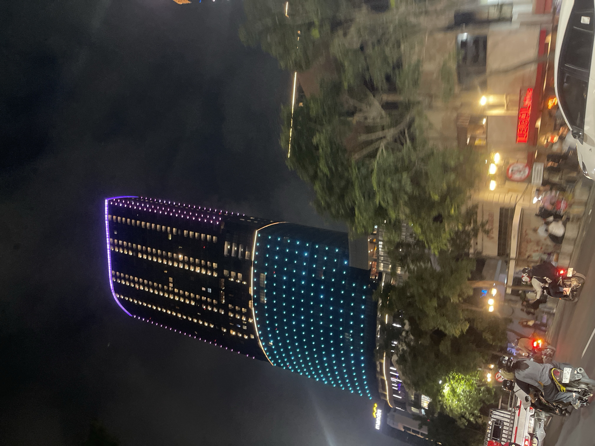 The image captures a nighttime street scene in Surabaya, Indonesia, dominated by the brightly illuminated Lumina Tower. The tower, a prominent landmark in the city, features a unique architectural design with its upper section adorned with purple lights and the lower curved section illuminated by a grid of vibrant blue lights. Below the tower, a bustling street is visible with several motorbikes carrying riders, indicating active traffic.
In the foreground, a white car is partially visible. The street is lined with trees and buildings, some of which are also lit up, contributing to the overall urban ambiance. People are seen on the sidewalks and near the motorbikes, suggesting a lively evening in the city. The overall impression is of a dynamic urban environment at night, showcasing a modern architectural marvel alongside the everyday activities of the city.
