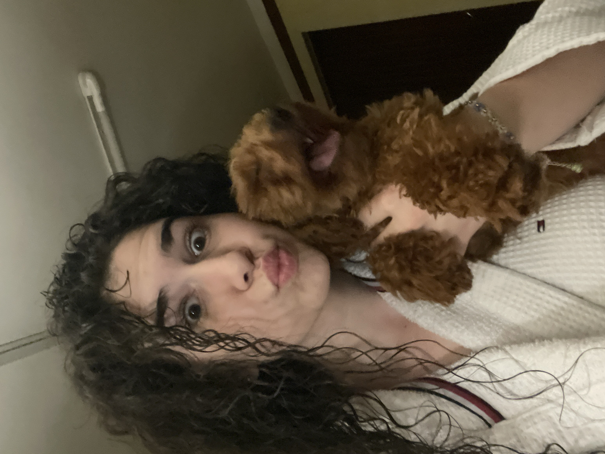 A person with dark curly hair is lying on their back, wearing a white textured robe. A small brown poodle is lying on the person's chest, facing right. The person has wide eyes and is making a kissing face. The background is blurry and dark, but a white vertical bar is visible on the left, and a dark rectangle is visible at the top. The location cannot be determined to be in the city of Sofia, Bulgaria based on the given image.