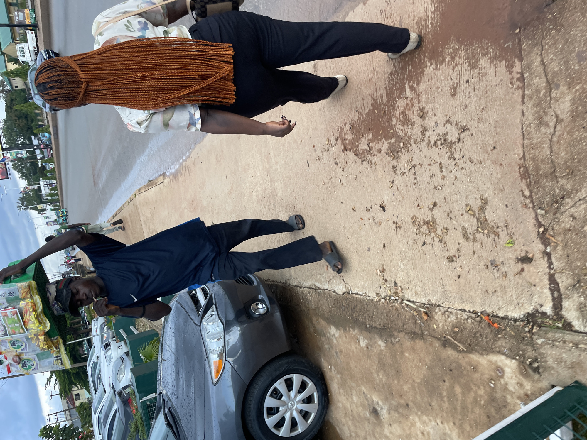 In Jos, Nigeria, a young man carrying a green tray of goods on his head walks along a wet, dirty sidewalk, smoking a cigarette. He wears a dark blue t-shirt and black trousers. A dark gray car is parked on the left side of the street. A woman with long, reddish-brown braided hair walks away from the camera on the same sidewalk. She is wearing a patterned shirt and black trousers. The street behind them has several cars and a few buildings in the distance. The sky is overcast.