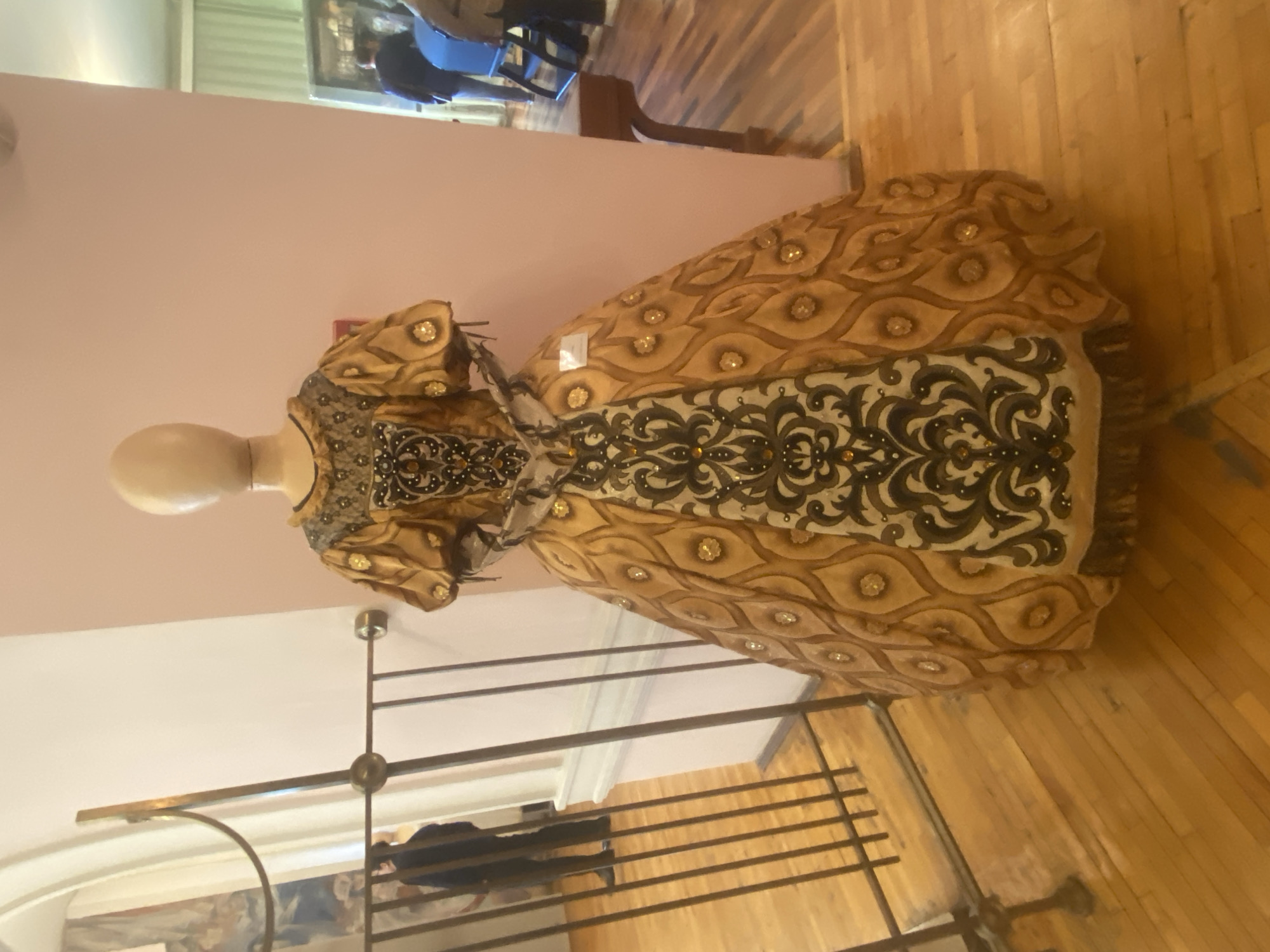 The image shows a mannequin wearing an ornate, golden dress with a large, black floral design on the bodice. The dress is on display behind a metal railing, likely in a museum or gallery. The background includes a pink wall, a glimpse of other people in the building, and a wooden floor. Based on the subject matter and architectural style, this could be in a historical building or museum in the city of Yekaterinburg, Russia.