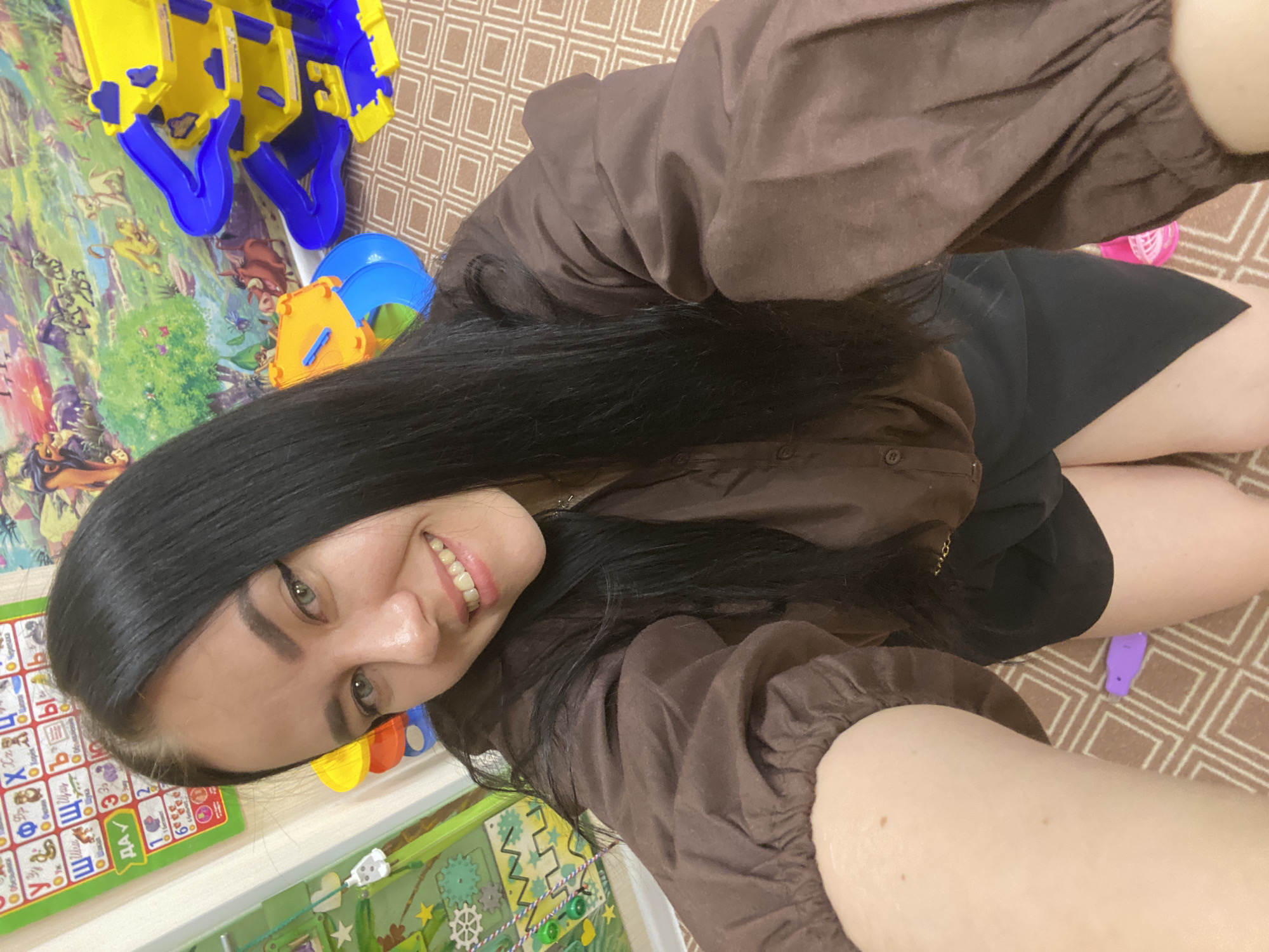 A woman is taking a selfie in what appears to be a child's playroom or bedroom. She is fair-skinned with long, dark hair and is wearing a brown blouse and a black skirt. Her pose suggests she is seated on the floor. The setting includes colorful children's toys and a rug with a geometric pattern. Based on the presence of Russian-language educational materials (alphabet chart), this photo may be in a location within Russia, specifically Chaikovsky, Russia.