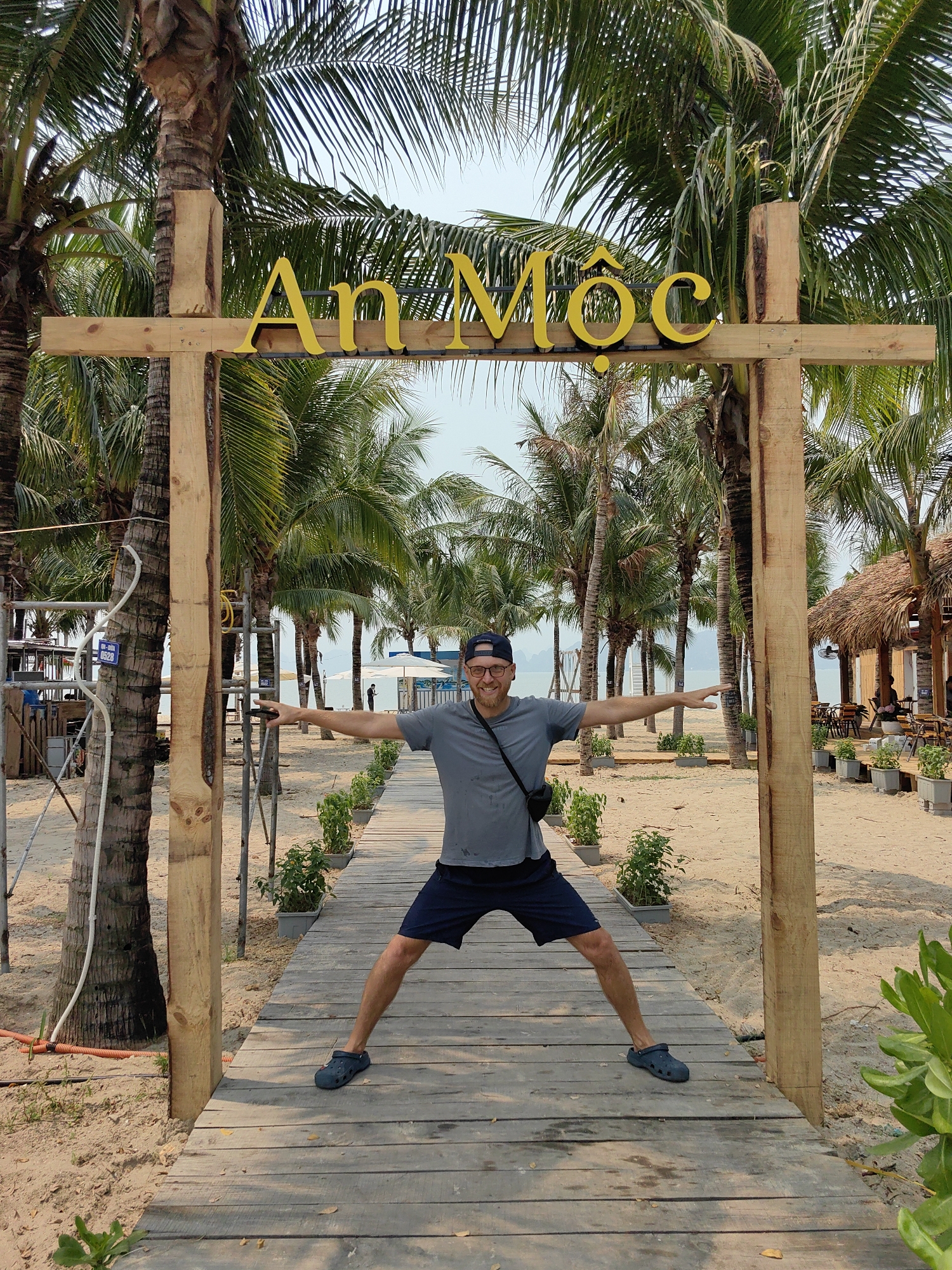A middle-aged man stands on a wooden boardwalk with his arms outstretched under a wooden sign. The sign has the words "An Mộc" in yellow. The man wears a gray shirt, blue shorts, a blue baseball cap, and blue Crocs. He also has glasses and a black messenger bag slung across his shoulder. The boardwalk leads to a beach. Behind the sign and boardwalk are palm trees. To the right is a thatched-roof structure with tables and chairs. The location is Bai Chay Ward, Vietnam.