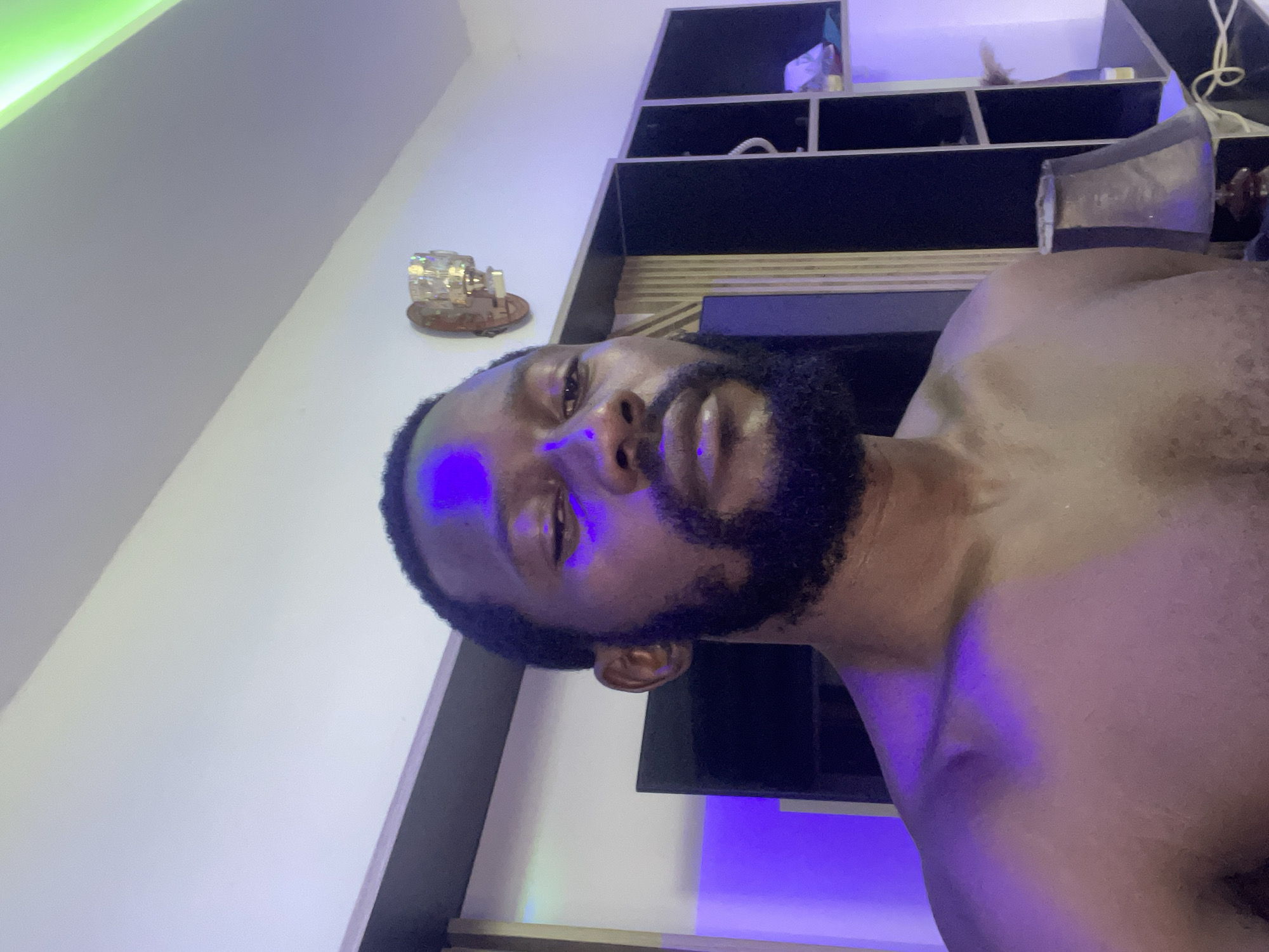 A shirtless male with a dark beard and short hair is the central subject of this indoor, vertically oriented image. He is looking directly at the camera with a neutral expression, his face partially illuminated by a strong blue/purple light emanating from below and to his left. His right cheek, jawline, and neck are also cast in this purplish glow.
The setting appears to be a modern room, likely at night or in low light, given the reliance on artificial illumination. In the background, to the subject's right, there are dark-colored shelving units, possibly built into a wall, with various indistinct items placed on them. Above the shelving, a bright green light source is visible, illuminating the upper part of the wall. To the subject's left, another strong blue/purple light source is present, complementing the one on his face and reflecting off the wall. A decorative object, possibly a small crystal lamp or ornament, sits on a shelf to the left of the subject's head.
The overall ambiance is dim with vibrant splashes of colored light (green, blue, and purple), creating a distinct mood. The image is rotated 90 degrees clockwise, presenting the subject horizontally across the frame. There is no visible text. The location context is Abuja, Nigeria.
