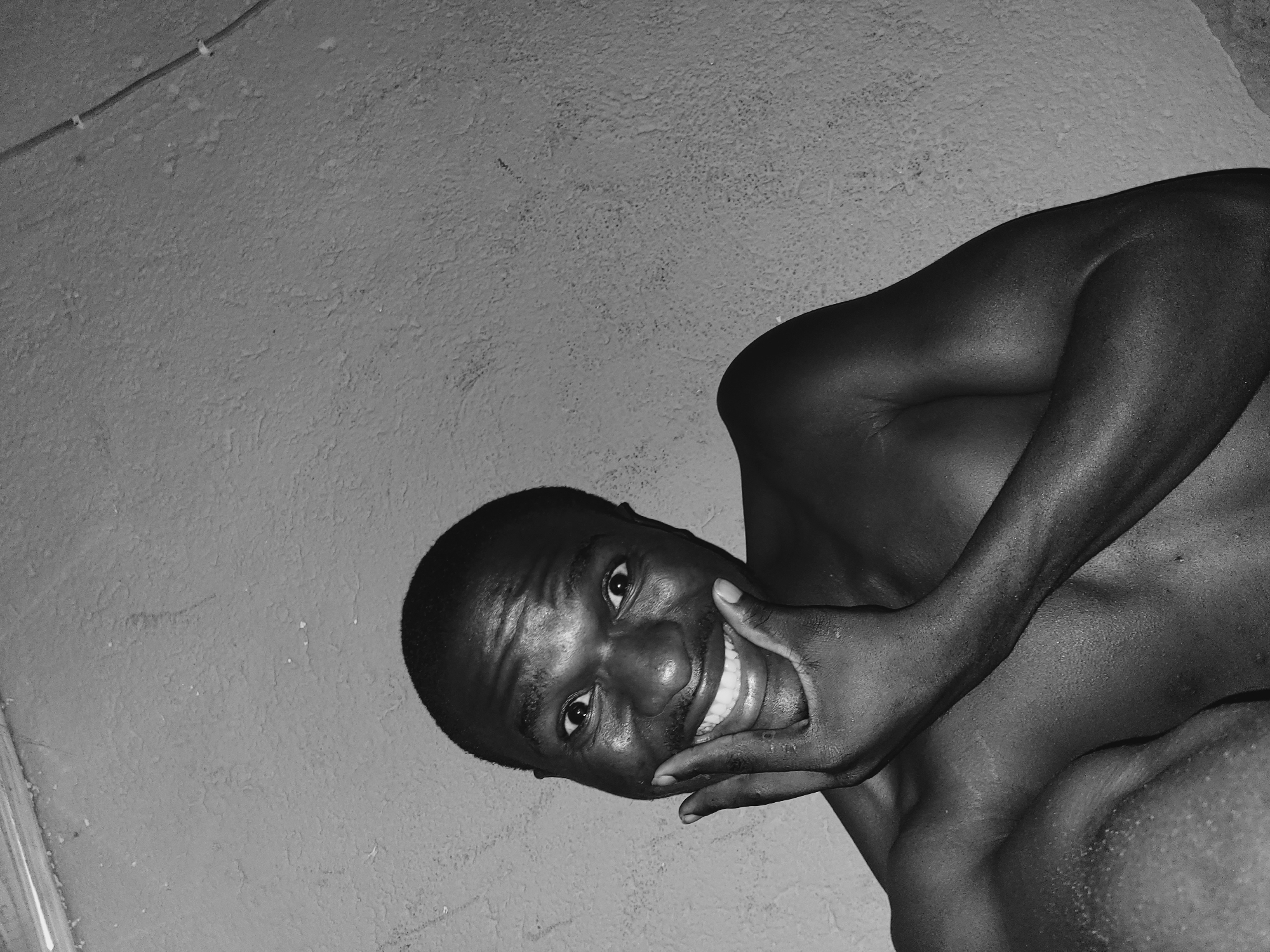 A black and white, close-up, waist-up photograph shows a shirtless Black man with short hair and a mustache. He is looking directly at the camera and smiling, with his right hand held to his chin. The background is a textured wall. The lighting is dim, casting shadows on the man's face and body. The image appears to have been taken indoors. There are no visible signs of the specific location in Bukuru, Nigeria, beyond the implied presence of an indoor setting with a wall.