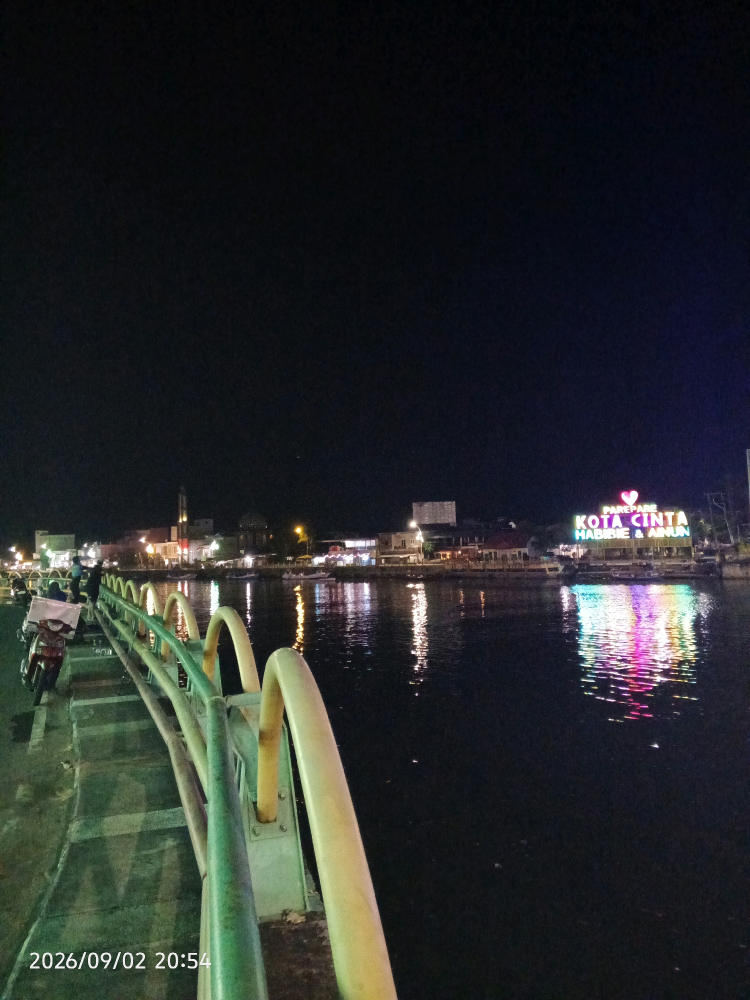 The image is a night-time view of a waterfront promenade in Parepare, Indonesia. In the foreground, a pedestrian walkway with a green and yellow railing stretches to the left. A motorcycle is parked on the walkway on the left side of the frame. In the background, across the dark water, is the city skyline illuminated by lights. A prominent illuminated sign on the waterfront reads "PAREPARE KOTA CINTA HABIBIE & AINUN" with a heart symbol above the word "CINTA". The lights from the city and the sign are reflected in the water. The sky is dark, indicating it is nighttime. A date and time stamp, "2026/09/02 20:54", is visible in the bottom left corner of the image.