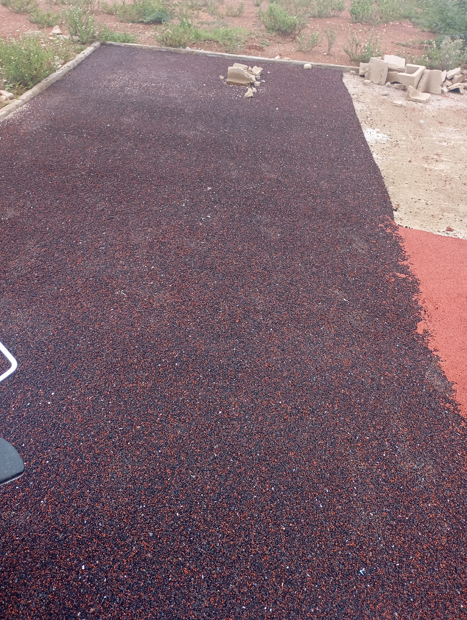 The image shows a newly laid surface, possibly a sports court or pathway, in an outdoor setting. The surface is composed of dark reddish-brown granules, appearing to be rubberized material, with some lighter flecks scattered throughout. The material is spread evenly across a section of what seems to be a prepared base, bordered by concrete edging on the left and top. In the background, there is dry, sparse vegetation and reddish-brown soil, indicative of a semi-arid or seasonally dry environment. To the right, a section of the surface is still unfinished or is a different material, appearing as a lighter beige-colored surface, possibly sand or unworked ground, with some scattered concrete debris. There is no clear indication of the city of Jos, Nigeria, within the visual information provided. A portion of a white metal railing or tool is visible in the lower left corner, and a dark object, possibly a piece of equipment or a shoe, is visible in the extreme lower left. The overall impression is of a construction or renovation project in progress.