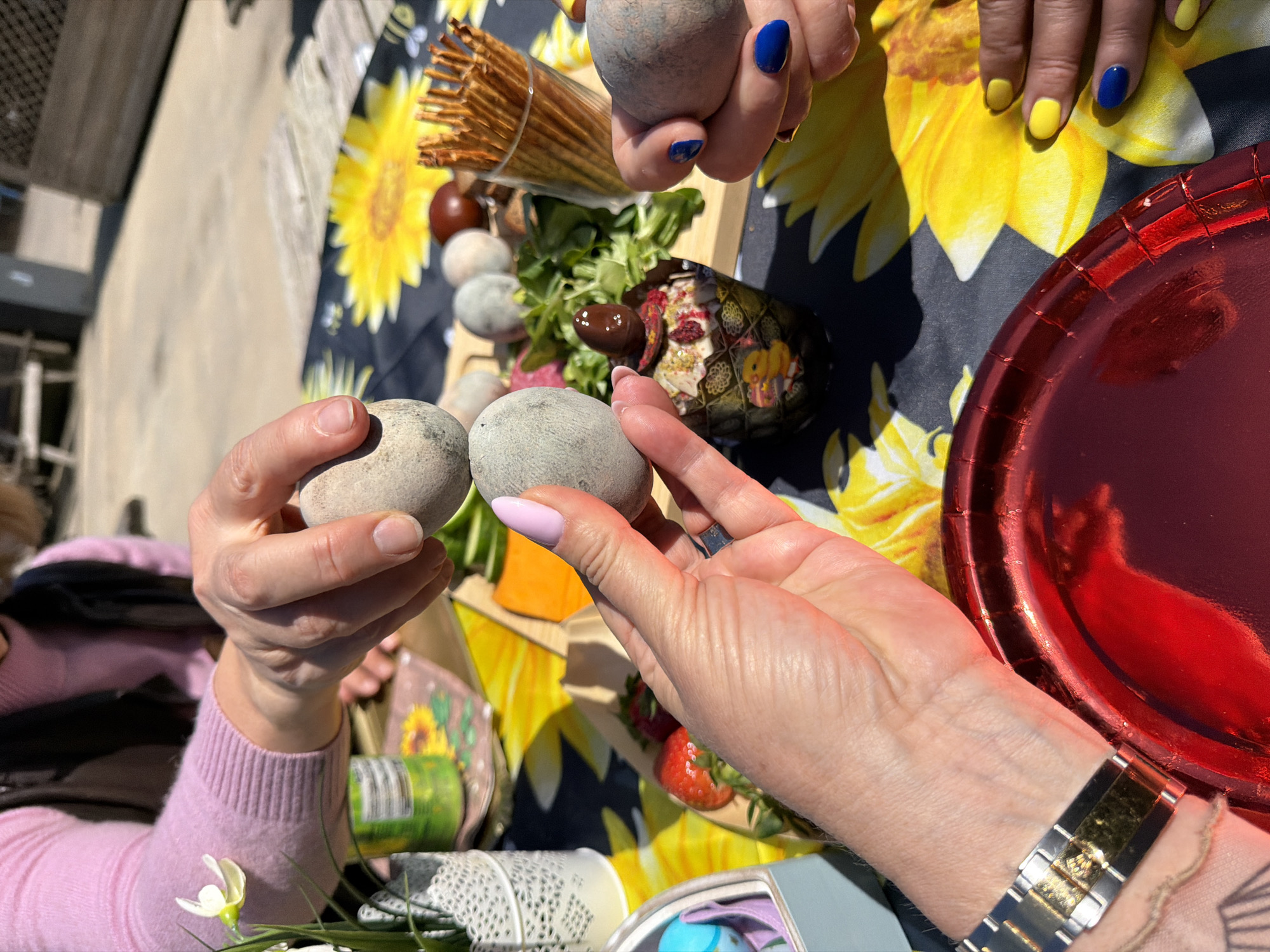 The image captures a festive outdoor gathering, likely an Easter celebration, set in Monistrol de Montserrat, Spain. The scene is viewed from above, focusing on hands and a table laden with food and decorations under bright sunshine.
Several hands are prominently featured, engaged in an egg-tapping game, a traditional Easter activity. In the foreground, two hands hold grey, speckled eggs, appearing to clink them together. One hand has light pink nail polish, while the other has natural nails. Another hand from the upper right, adorned with vibrant blue nail polish, also holds a similar grey egg. Further back, another pair of hands displays an alternating pattern of bright yellow and blue nail polish.
The table is covered with a dark blue cloth featuring large, cheerful yellow sunflower patterns, enhancing the spring and festive atmosphere. Various items are spread across the table: a glass filled with thin breadsticks, small round fruits or olives, fresh green lettuce, and slices of orange food, possibly cheese or fruit, arranged on wooden serving boards. A decorative egg-shaped bowl, intricately patterned, holds a dip or salad garnished with what looks like dried berries. A shiny red metallic plate is visible on the right side of the frame, along with a white lace-patterned cup or holder and a small white flower with a green stem, adding to the decorative elements.
A person wearing a light pink long-sleeved top is visible on the left. On the right, a wrist adorned with a silver and gold watch is seen. The strong sunlight casts distinct shadows, indicating a bright daytime setting. The overall mood conveyed is one of joy, tradition, and communal celebration in an inviting outdoor environment. No clearly legible text is visible on any objects in the image, though a barcode can be seen on a green can in the background.