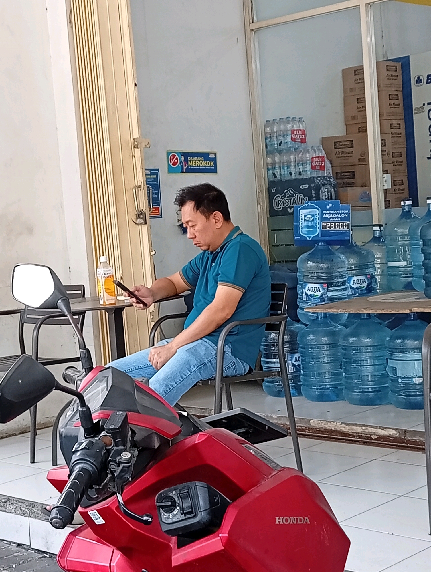 A middle-aged East Asian man sits on a chair next to a red motorcycle, looking at his smartphone. He is wearing a teal polo shirt and blue jeans. He is seated at a small table with a bottle of orange drink on it. In the background, there is a store with shelves stocked with bottled water and cardboard boxes. A "No Smoking" sign is visible on the wall. The scene appears to be outdoors, possibly in front of a shop. The location is identified as Surabaya, Indonesia.