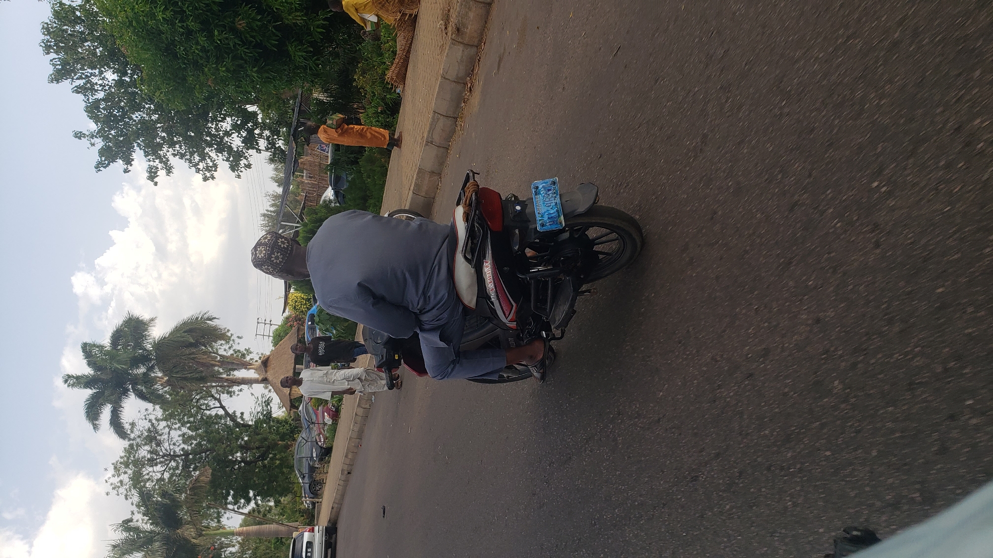 A person wearing a gray outfit and a patterned cap is riding a motorcycle on a paved road. The motorcycle has a blue license plate. On the left side of the road, several people are walking along the side of the road. A tall palm tree and other greenery are visible. Cars are parked behind the people. On the right side of the road, there are more trees and shrubs. The road appears to be in Hayin Banki, Nigeria.