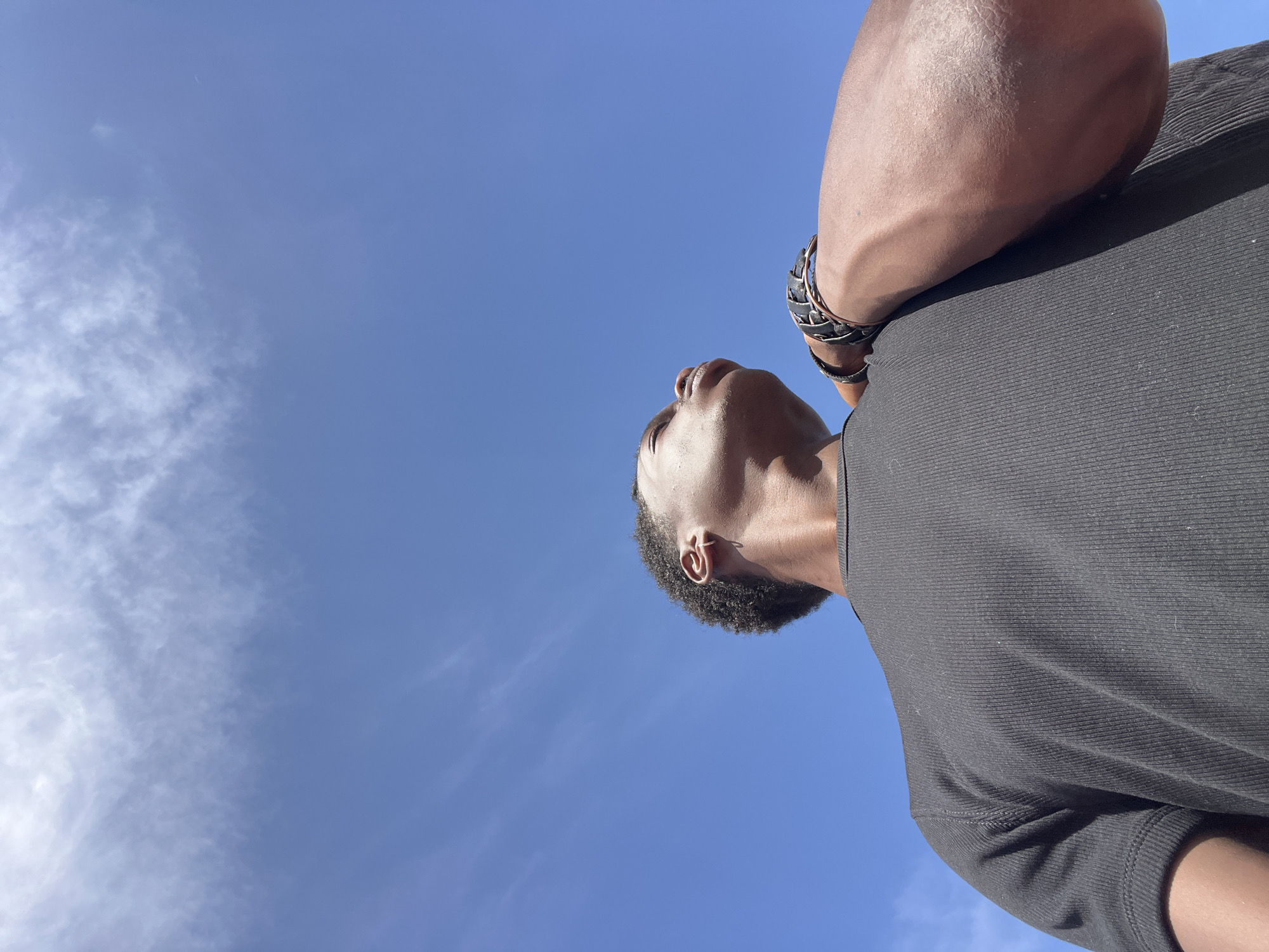 A low-angle, waist-up shot shows a person looking upwards against a bright blue sky with scattered white clouds. The person, viewed from the chest up, is wearing a black, ribbed t-shirt. Their right arm is bent, with their hand resting on their shoulder, revealing a black and silver braided bracelet on their wrist. The skin tone is dark. The image was taken in Sant Adrià de Besòs, Spain.