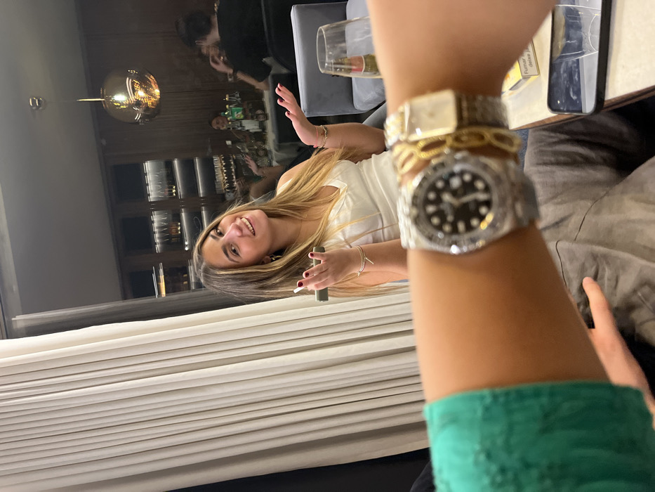 An indoor scene depicts a social gathering, likely within an upscale restaurant or bar. The specific location within Lordelo do Ouro e Massarelos, Portugal, cannot be determined from visual evidence.
In the foreground, a person's left arm, wearing a green sleeve, is positioned prominently, displaying a silver-toned wristwatch with a black dial and decorated bezel, a gold-toned chain-link bracelet, and a second gold-toned rectangular wristwatch. The arm is holding a smartphone, suggesting it belongs to the photographer. On a light-colored table to the right, a white and yellow cigarette package with the text "FUMAR" is visible, along with a dark smartphone and a clear glass.
The mid-ground features a young woman with long, light brown hair, smiling directly at the camera. She wears a white v-neck top and multiple thin bracelets on both wrists. Her left hand, with red painted fingernails, holds a dark rectangular object, possibly a vape pen or small device. Her right hand is open, palm up, in a gestural pose.
The background is dimly lit and out of focus, revealing several other patrons seated at tables. Decorative elements include tall, light-colored pleated curtains to the left, a gold-toned spherical pendant light fixture, and a dark wood shelving unit or wall paneling illuminated by strip lighting, possibly displaying bottles. Grey upholstered seating and tables with light surfaces are visible. The overall environment suggests a modern, relaxed, indoor social setting.