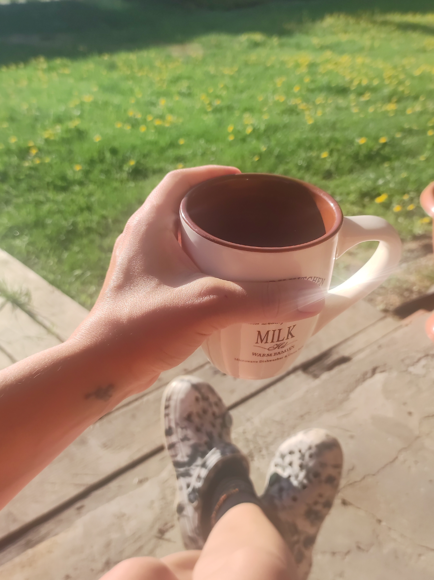 A person holds a white mug with brown trim in their hand, with the words "MILK" and "WARM FAMILY" printed on the side. The mug is empty. The person is wearing spotted Crocs. The background is a green lawn with yellow wildflowers. The surface the person is sitting on appears to be a wooden deck. The location is impossible to determine.