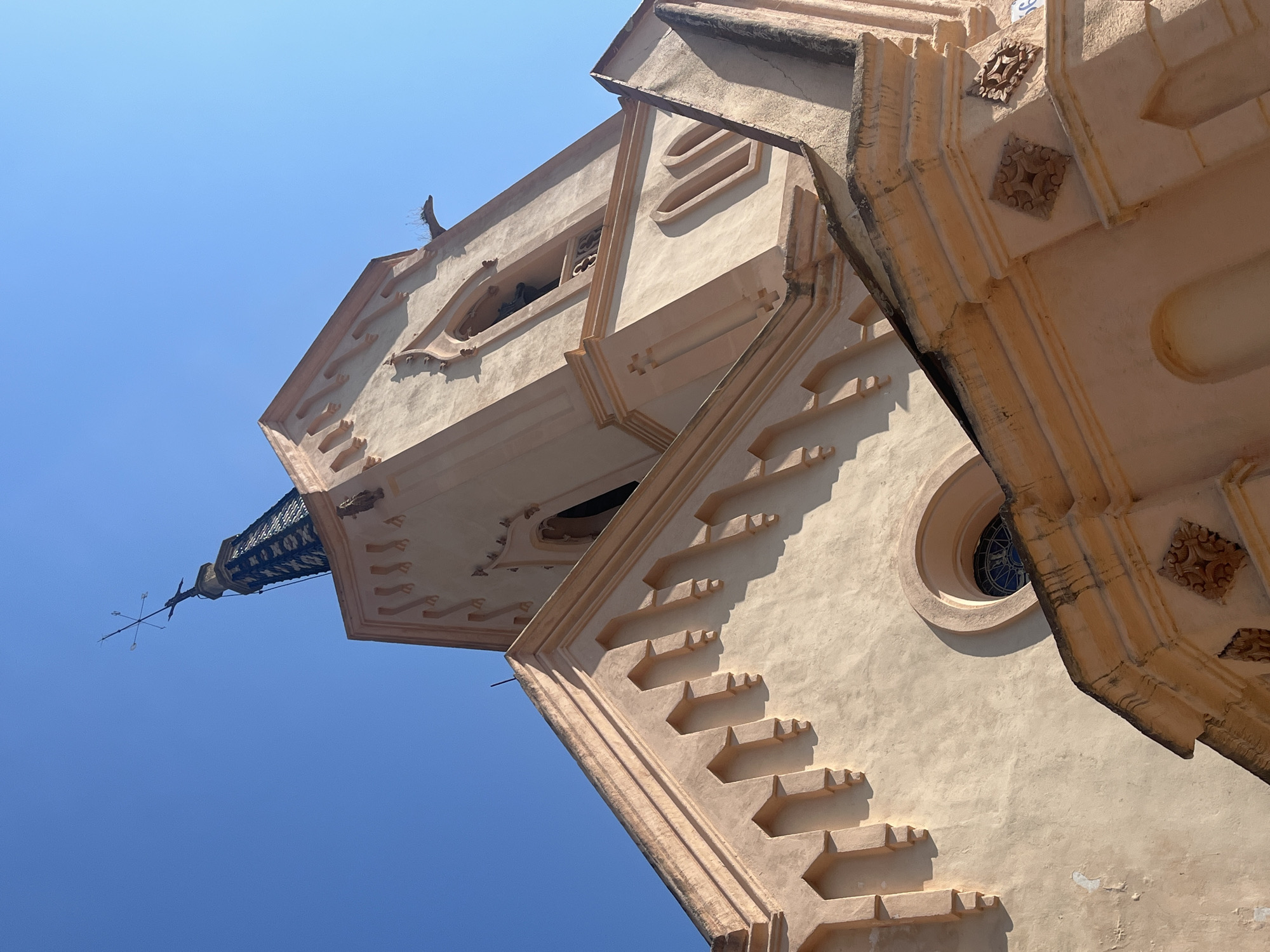 Looking up at a tall, light-tan building with intricate architectural details against a bright blue sky. The building features decorative moldings, geometric patterns, and a small, round window. At the top, a weather vane is visible. The location is Unknown, Unknown.