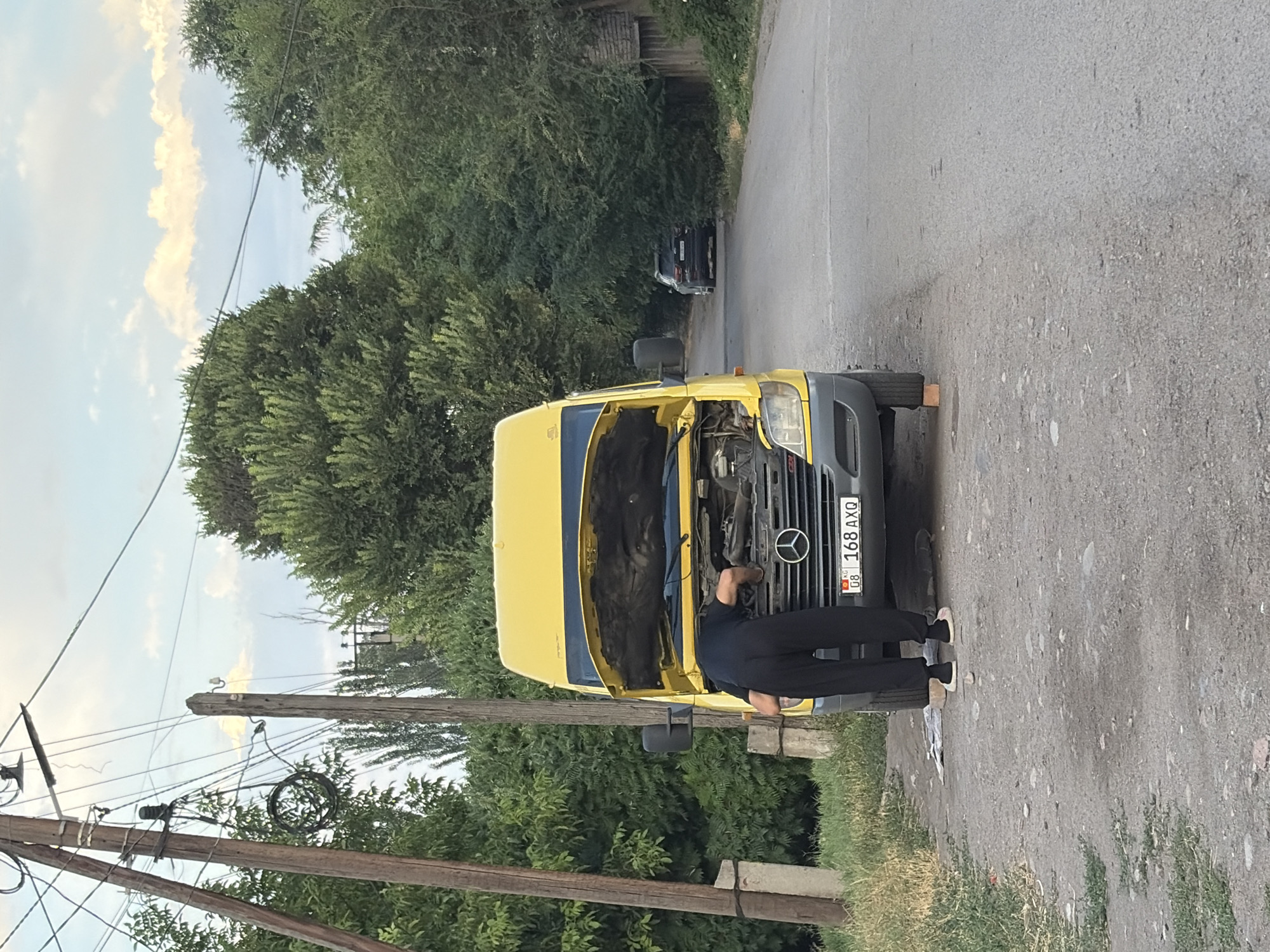 A person is working on the engine of a yellow Mercedes-Benz Sprinter van. The van's hood is open, and the person is bent over, appearing to be inspecting or repairing the engine. The license plate of the van is visible and reads "KG 168 AXQ". The scene takes place on a paved road in a somewhat rural or suburban area, with trees and vegetation lining the sides. In the background, there is a dark-colored SUV parked on the side of the road and a wooden building. Utility poles with wires are prominent in the left foreground, suggesting the presence of infrastructure. The lighting suggests it is daytime, possibly late afternoon, given the soft light and the presence of shadows. The ground is a mix of asphalt and gravel. The general environment suggests a location outside of a dense urban center, potentially on the outskirts of the city of Kant, Kyrgyzstan.