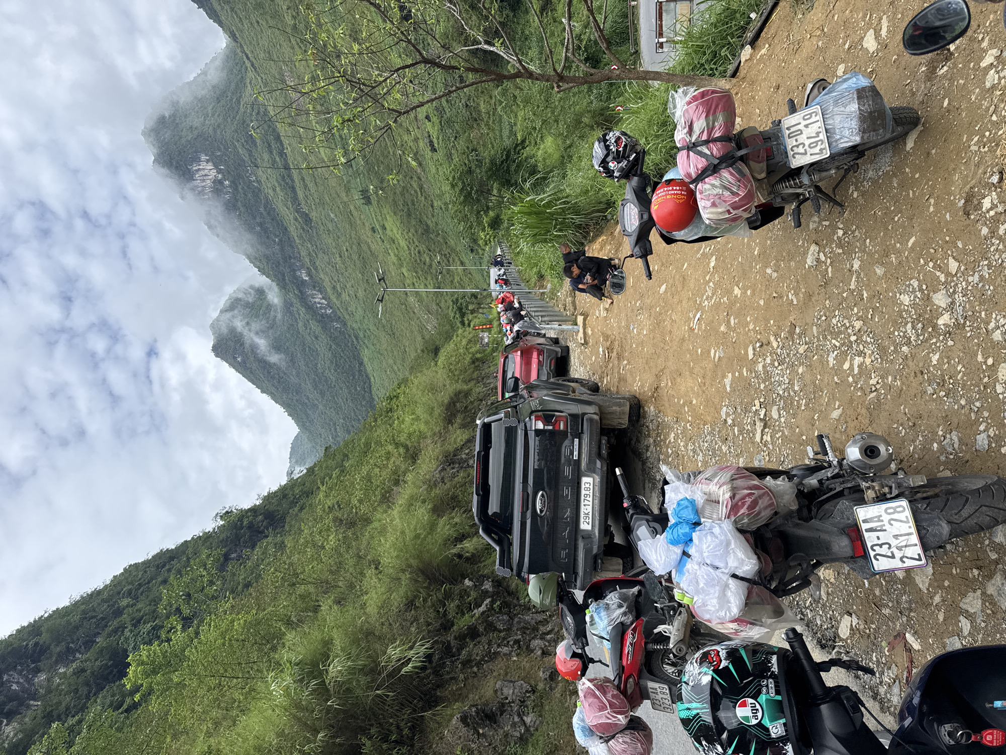 A line of parked vehicles, primarily motorcycles heavily laden with luggage, and a dark-colored pickup truck, are oriented along a dirt road. The vehicles are positioned roadside to the right, with a steep, verdant mountain rising to the left of the frame. Overcast skies and mist-shrouded mountain peaks are in the background. The license plates on the vehicles indicate that the location is likely Yên Minh Commune, Vietnam. People are seen standing and walking along the road between the vehicles.