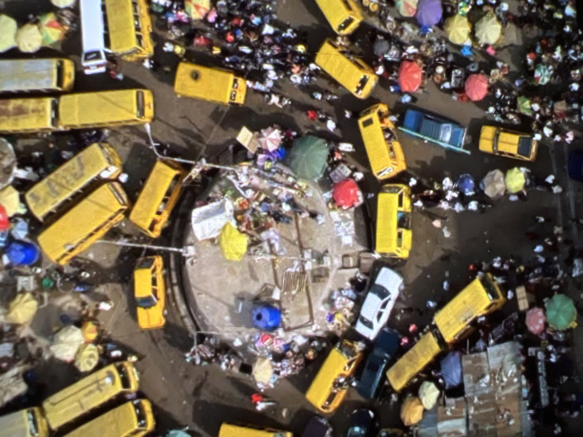 This aerial image depicts a bustling, crowded marketplace in Lagos, Nigeria, likely a motor park or a major transport hub. The scene is dominated by a multitude of yellow minibusses, known locally as "danfos," which are the primary mode of public transportation in the city. These buses are parked closely together, some appearing to be loading or unloading passengers. Surrounding the buses, a large number of people are visible, moving about the area, interacting with vendors, or waiting for transportation.
The ground is a mix of paved and unpaved surfaces, with scattered debris and refuse indicating the high level of activity and the informal nature of the market. Numerous umbrellas, in various colors and sizes, are deployed by vendors to shade their goods or by individuals to protect themselves from the sun or potential rain. These umbrellas, along with colorful fabrics and stalls, add vibrant splashes of color to the otherwise muted tones of the buses and the ground.
In the background, some structures suggest the presence of buildings or makeshift shelters, typical of a busy urban market environment. The overall impression is one of intense activity, movement, and a dense concentration of people and vehicles. The image captures the energetic and chaotic rhythm of daily life in a Nigerian city.