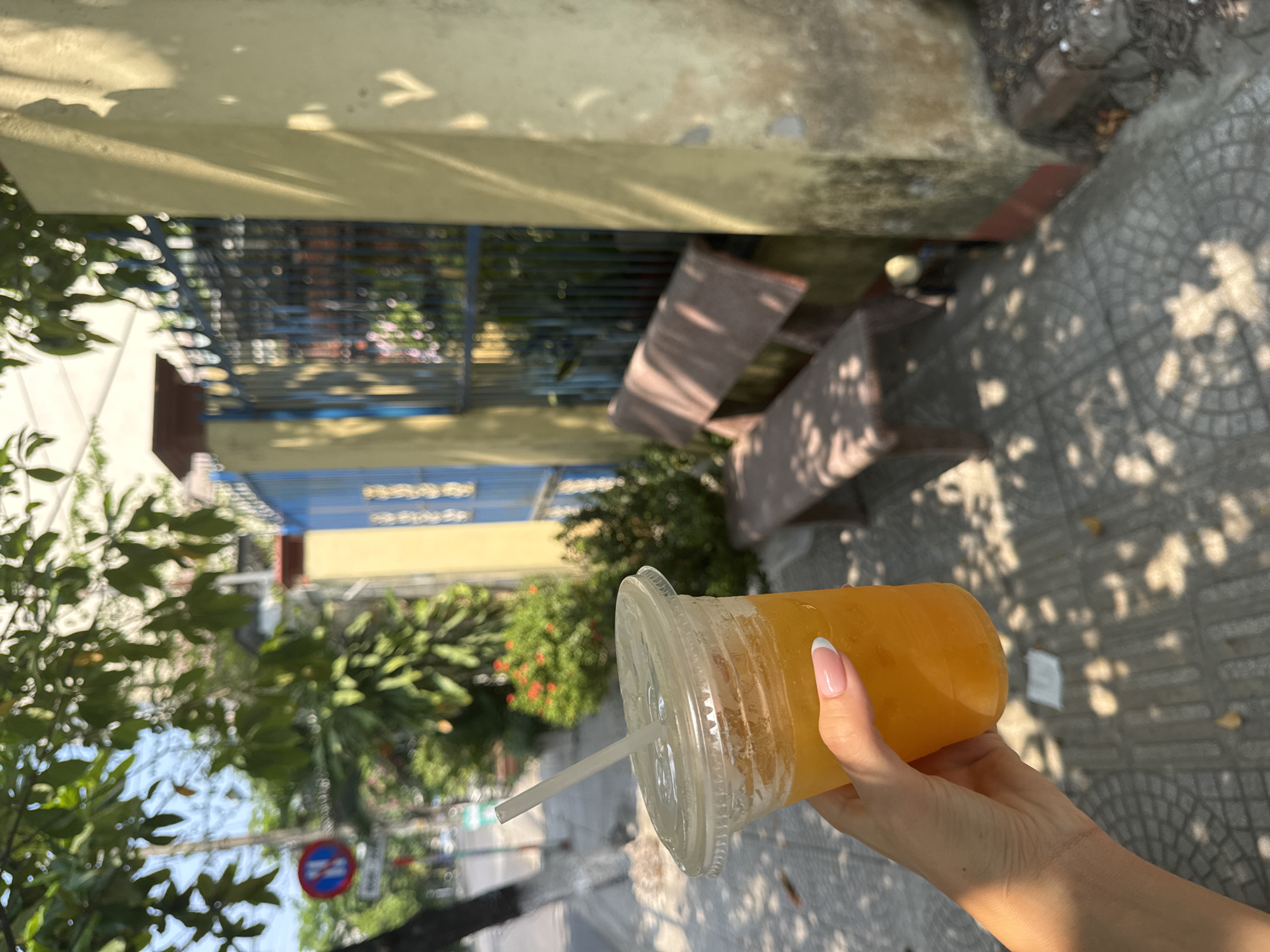 A woman's hand, with a French manicure, holds a plastic cup filled with an orange-colored beverage and a straw. The photograph appears to have been taken outdoors, likely on a sidewalk in Đà Nẵng, Vietnam, based on the architecture and vegetation visible in the background. A tree is partially obscuring the left of the shot. A building with blue and yellow elements is visible behind the tree. A pedestrian walkway is visible on the right of the hand and drink. There are also brown-colored seats nearby.