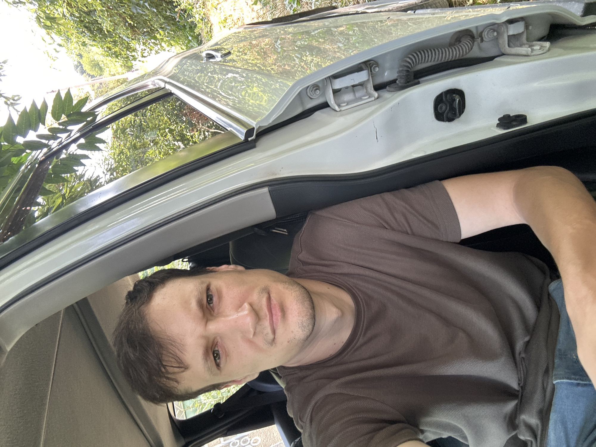 A man is sitting in the driver's seat of a light-colored car, with the driver's side door open. He is looking at the camera and has short brown hair and light skin. He is wearing a brown t-shirt and blue jeans. The car appears to be parked on a dirt road, with lush green foliage and trees visible in the background. The lighting suggests it is daytime. The image was taken in Makiv, Ukraine.
