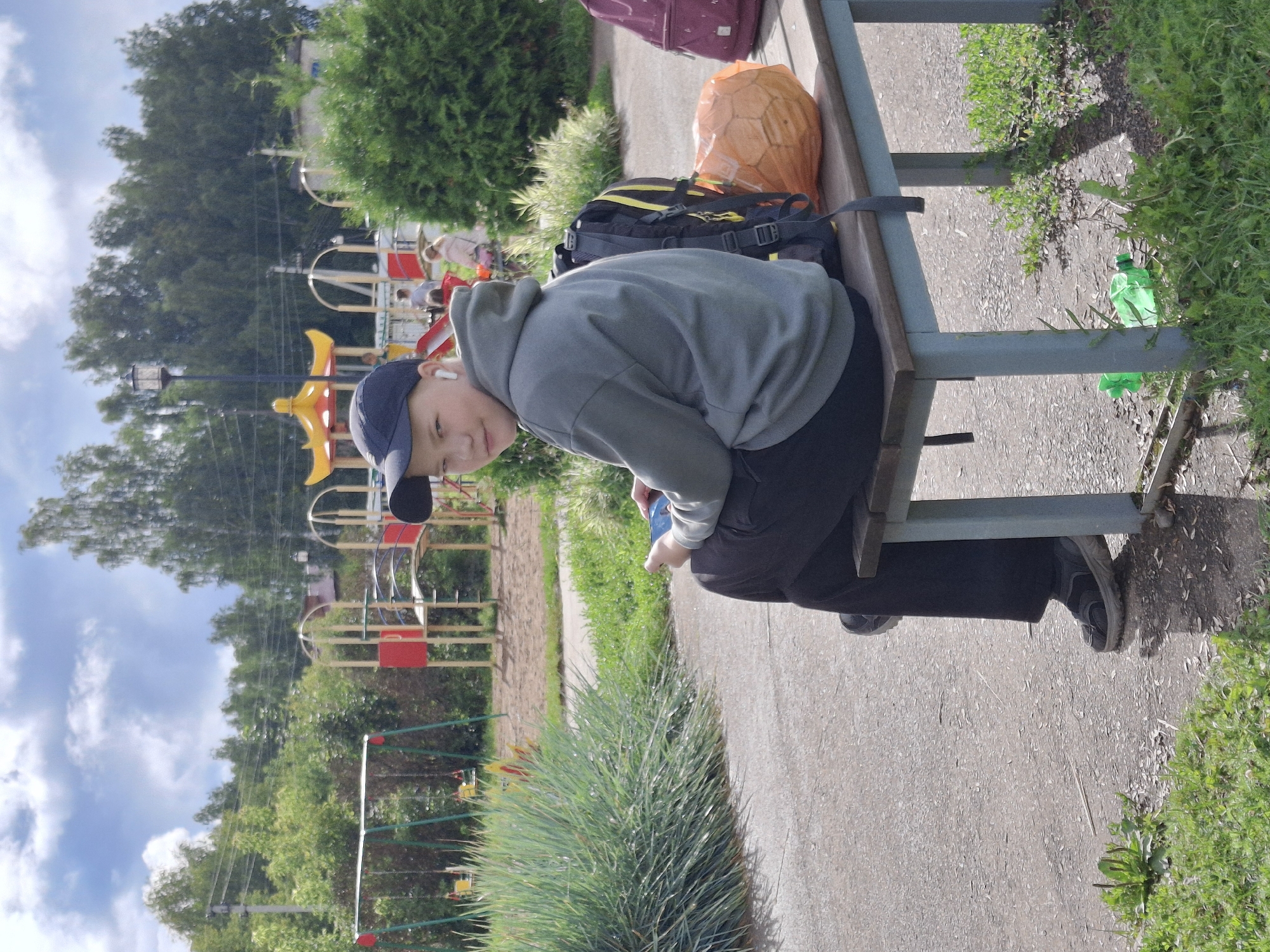 An outdoor medium shot shows a young person with light skin, wearing a dark blue baseball cap, a grey hooded sweatshirt, and dark pants, sitting on a wooden bench. They are holding a mobile phone in their right hand and wearing white wireless earbuds. A black backpack with yellow accents is on the bench to their left, and an orange ball is to the right of the backpack. In the background, a playground with swings, slides, and other play structures is visible, along with trees and a cloudy sky. The location is implied to be the city of Elovo, Russia.