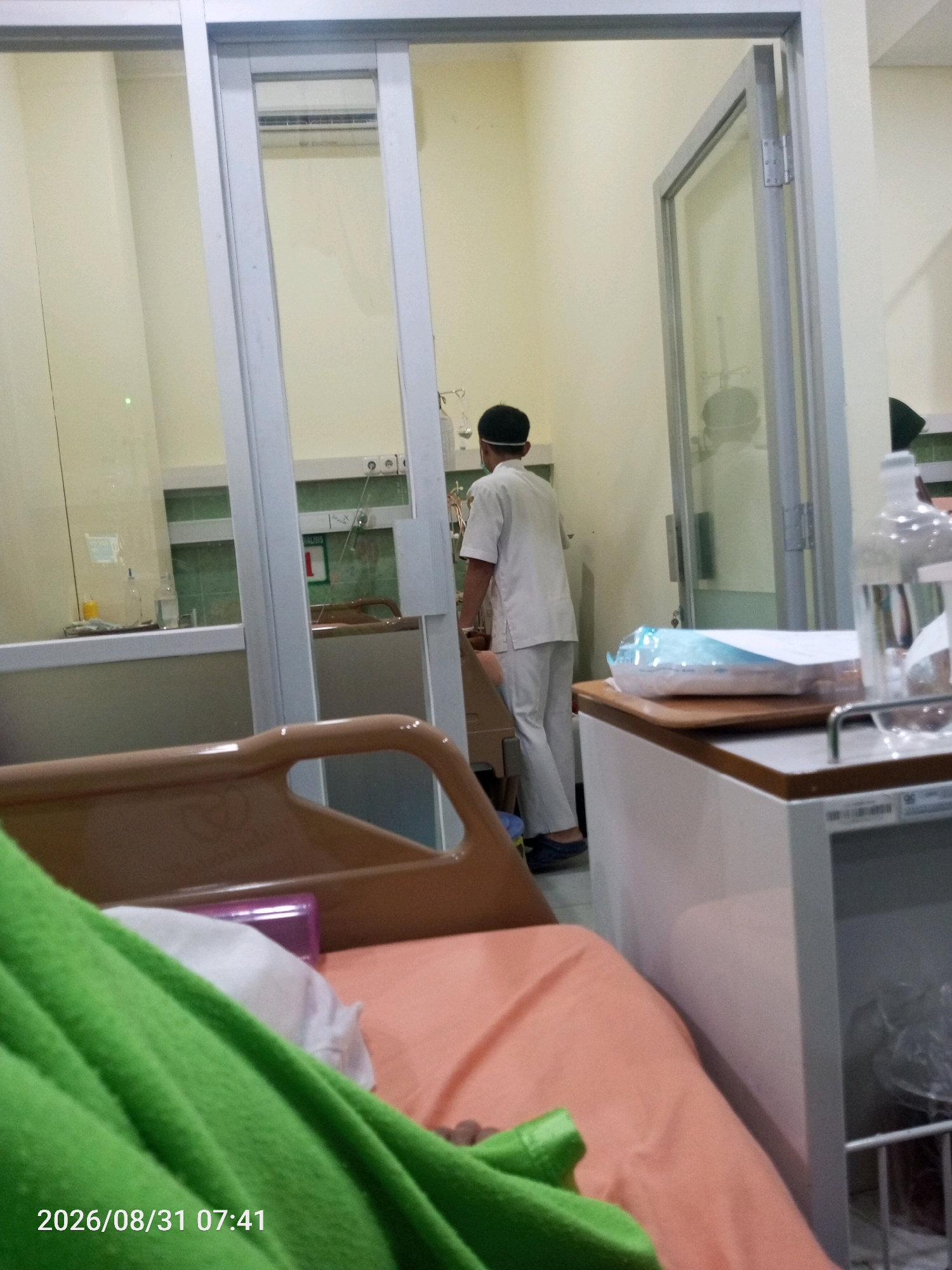 An interior scene, likely a hospital room in Banjarnegara, Indonesia, is depicted. A person wearing a white scrub suit and a dark cap is visible from the back, standing inside a room with a glass door that is partially open. They appear to be interacting with equipment or providing care. In the foreground, a patient is partially covered by a bright green blanket, resting on a pink bed. Medical equipment, including an IV stand and bottles, are present. The room is well-lit, with a sliding glass door on the left side and a standard door on the right. A date and timestamp, "2026/08/31 07:41," is overlaid on the image.