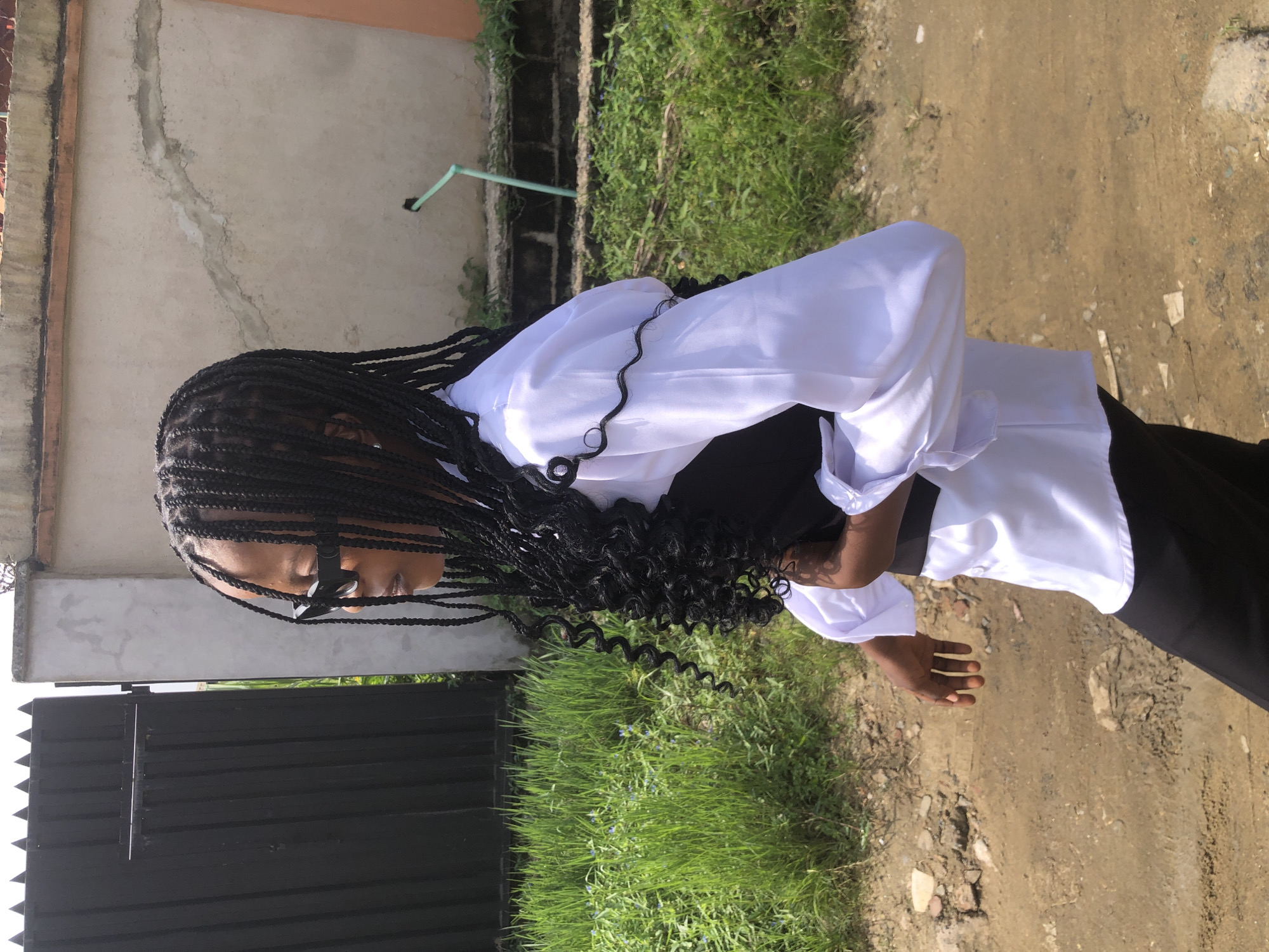 A young female with braided hair and sunglasses walks outdoors in Ado, Nigeria. She is wearing a white collared shirt, a black vest, and black pants. The background features a black metal gate, tall green grass, a light-colored concrete wall with some cracks, and a dirt path. The lighting suggests it is daytime.