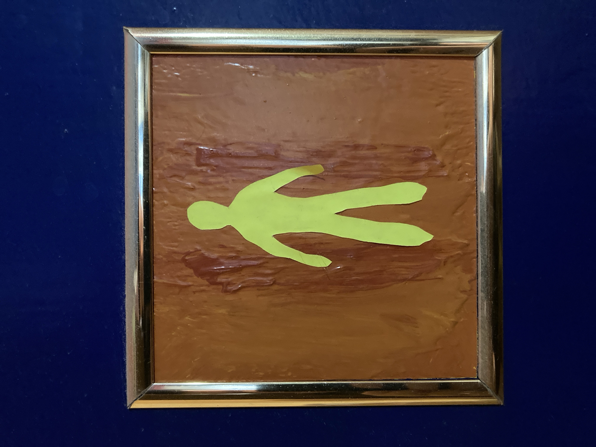 A framed, light green paper cutout of a person stands in the center of a framed, brown, textured surface. The frame is gold. The framed artwork is mounted on a dark blue background. The image is taken from a slightly angled perspective, with the top of the frame appearing closer to the viewer than the bottom. The lighting is even and does not cast strong shadows.