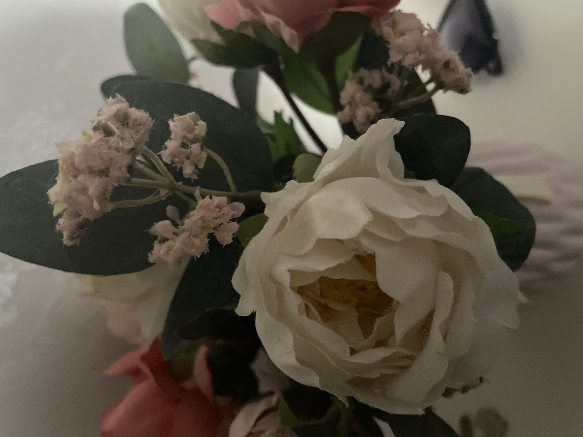 This media file is a close-up, slightly blurry photograph of a bouquet of artificial flowers. The primary subjects are the flowers themselves, which appear to be a mix of roses and smaller, delicate blossoms. One large, white rose is prominently featured in the foreground, its petals unfurling. To the left, a cluster of small, light pink flowers are visible, with darker green leaves providing a backdrop. In the upper part of the image, a pink flower can be seen, partially out of focus. The lighting is dim, suggesting it might be evening or an indoor setting with low light. The background is a soft, neutral color, possibly a wall or fabric, with some texture. There are no people or discernible activities taking place; the focus is solely on the floral arrangement. The overall impression is one of soft, muted colors and a gentle, perhaps serene, mood. There is no visible text in the image. The provided location context of Moscow, Russia, does not offer specific visual cues within the image itself.