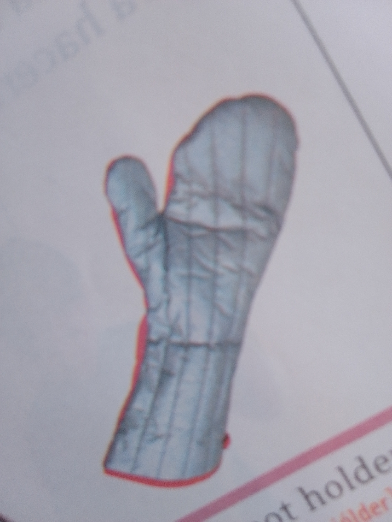 The media file shows a close-up, slightly blurred image of a single oven mitt. The mitt is light blue with gray stitching forming a quilted pattern. It has a red trim around the cuff. The mitt is depicted in a vertical orientation, with the thumb pointing towards the top left of the frame. The background is a light-colored surface with faint text, possibly from a book or magazine, visible in the upper left quadrant. A diagonal line in the upper right quadrant and some text at the bottom right of the frame suggest the image might be part of a larger layout or publication. The lighting appears to be neutral, with no strong shadows. There are no people or other objects clearly visible in the frame, and no specific time of day or weather conditions can be inferred. The context provided indicates the location is La Tagua, Venezuela. The text "ot holder" is partially visible at the bottom right of the image.